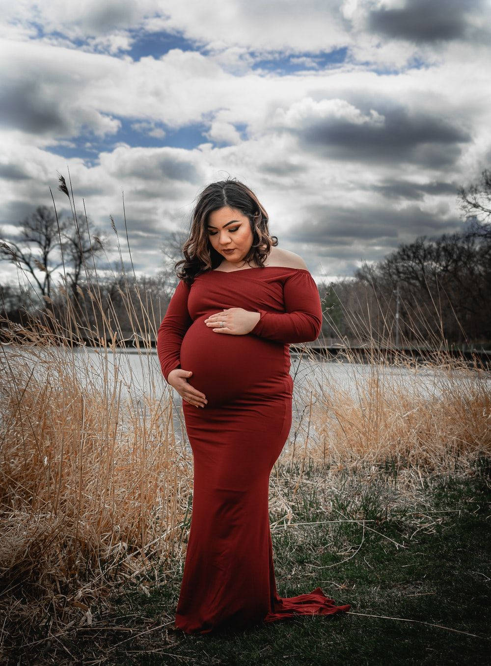 Pregnant 桌布 壁紙 — 一位身穿飄逸紅裙的孕婦在戲劇性的多雲天空下，在湖邊輕輕地抱著她的肚子。