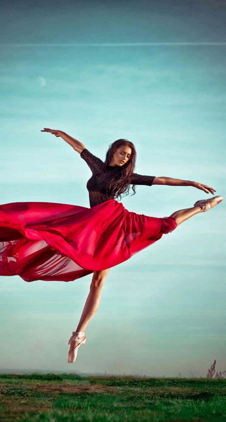 Ballet 桌布 壁紙 — 一位身穿飄逸紅裙的芭蕾舞者在草地上方柔和的青綠色天空背景下優雅地跳躍。