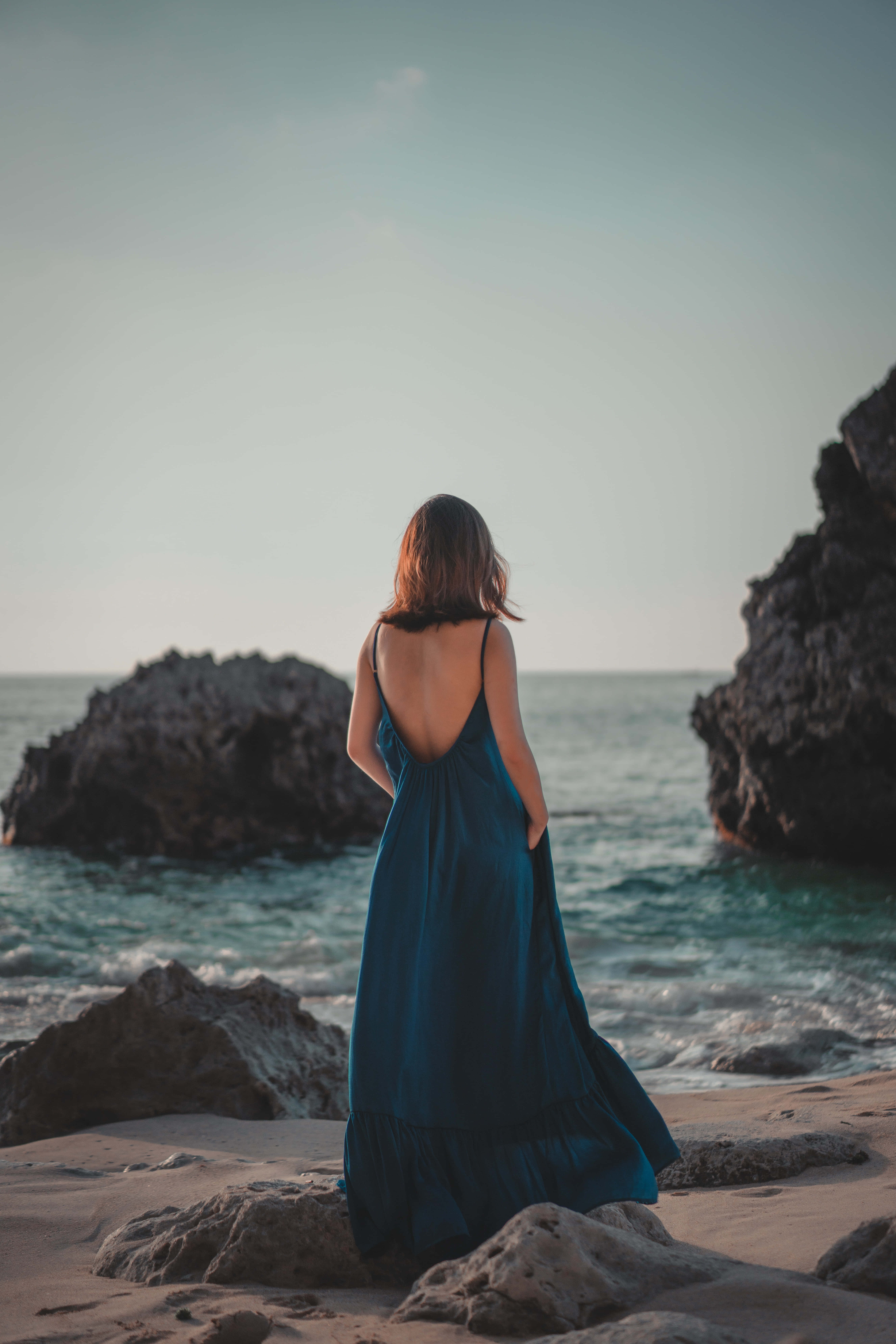 Navy Blue Dress 桌布 壁紙 — 一位身穿飄逸藍色連衣裙的女性站在日落時分的岩石海灘上，凝視著海洋。