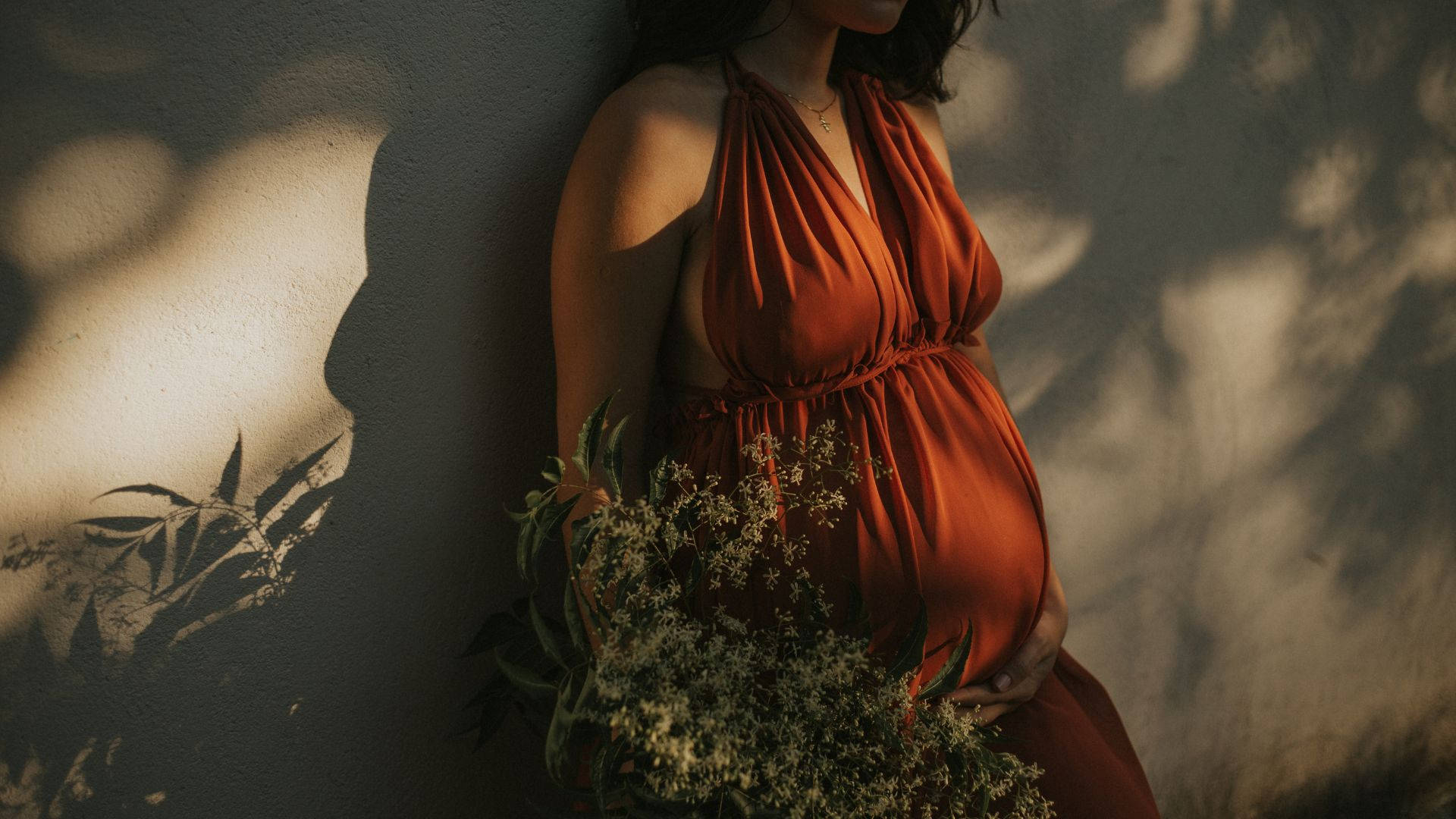 Woman 桌布 壁紙 — 一位身穿飄逸鏽色連衣裙的孕婦靠在陽光照耀的牆上，手持野花，斑駁的影子在她周圍舞動。