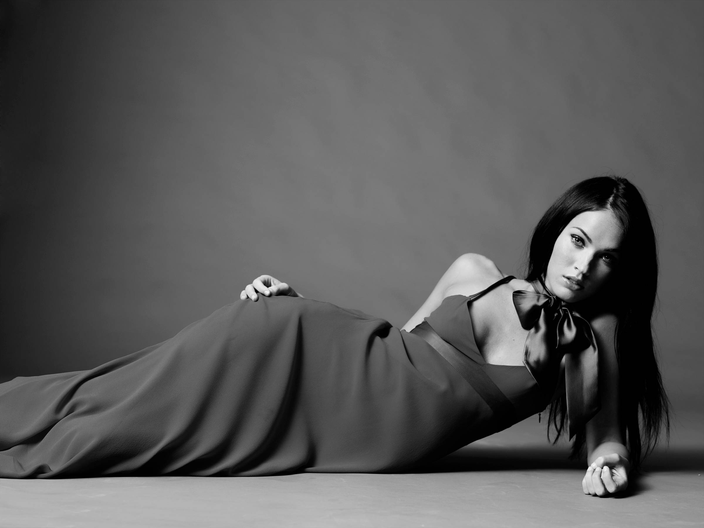 Megan Fox HD 桌布 壁紙 — 一位身穿飄逸長裙的女性躺在純色背景的地板上，以單色調的從容表情直視前方。