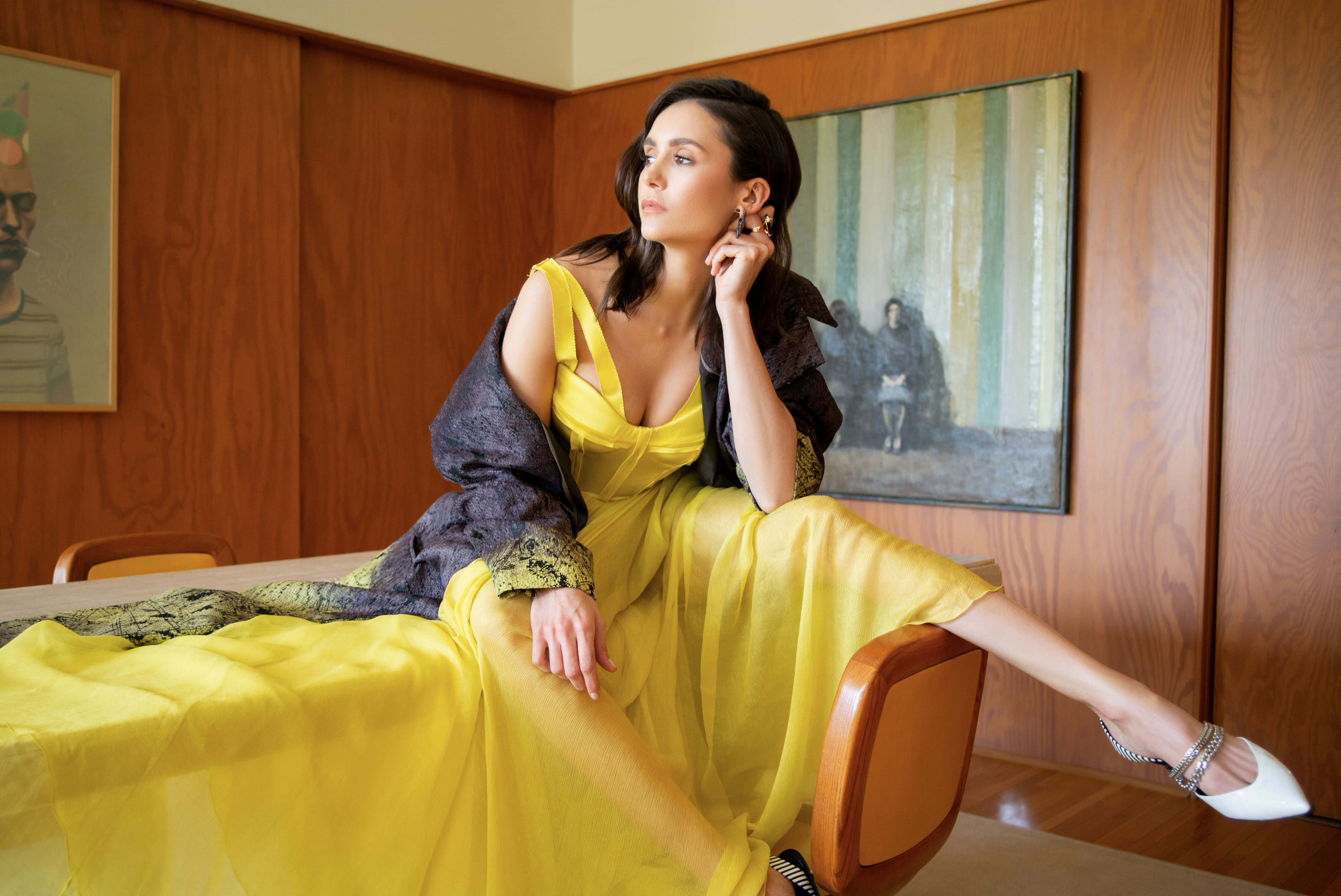 Yellow Dress 桌布 壁紙 — 一位身穿飄逸黃色連衣裙的女性，在溫暖燈光的中世紀現代房間裡，優雅地靠在木椅上。