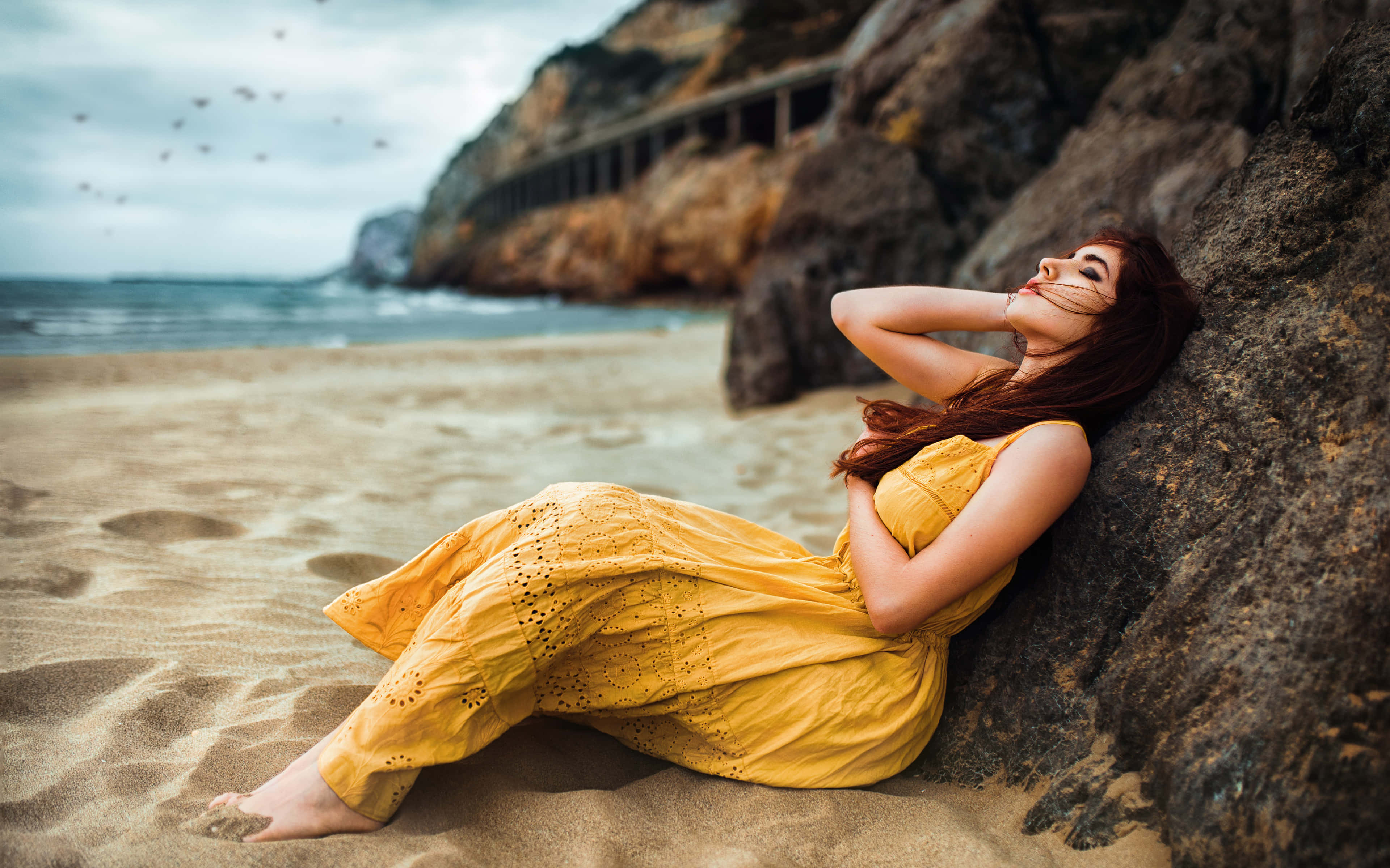 Beach Photography 桌布 壁紙 — 一位身穿飄逸黃色連衣裙的女性躺在沙灘上，靠著一塊大岩石，仰望天空，鳥兒在海洋附近飛翔。