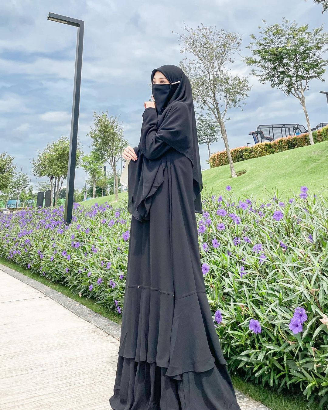 muslim 桌布 壁紙 — 一位身穿飄逸黑色長袍和頭巾的女性，站在多雲天空下充滿活力的紫色花壇旁。