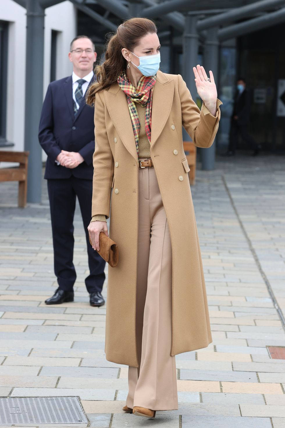 Kate Middleton 桌布 壁紙 — 一位身穿駝色大衣和格子圍巾的女性在鋪砌的戶外小徑上行走時揮手，身後有一位穿西裝的男士。