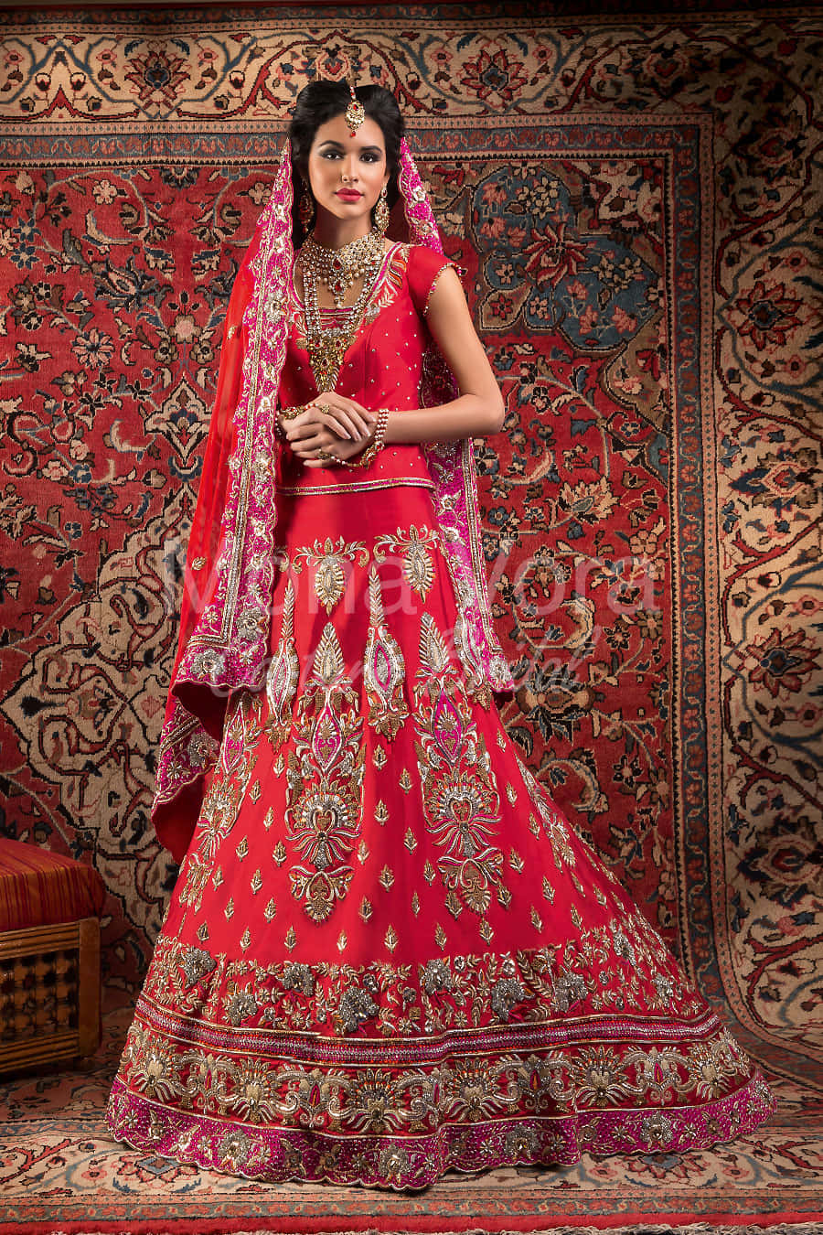 紅色動圖 桌布 壁紙 — 一位身穿鮮紅色刺繡 lehenga 並飾有金色細節的女性，優雅地站在華麗圖案地毯前。