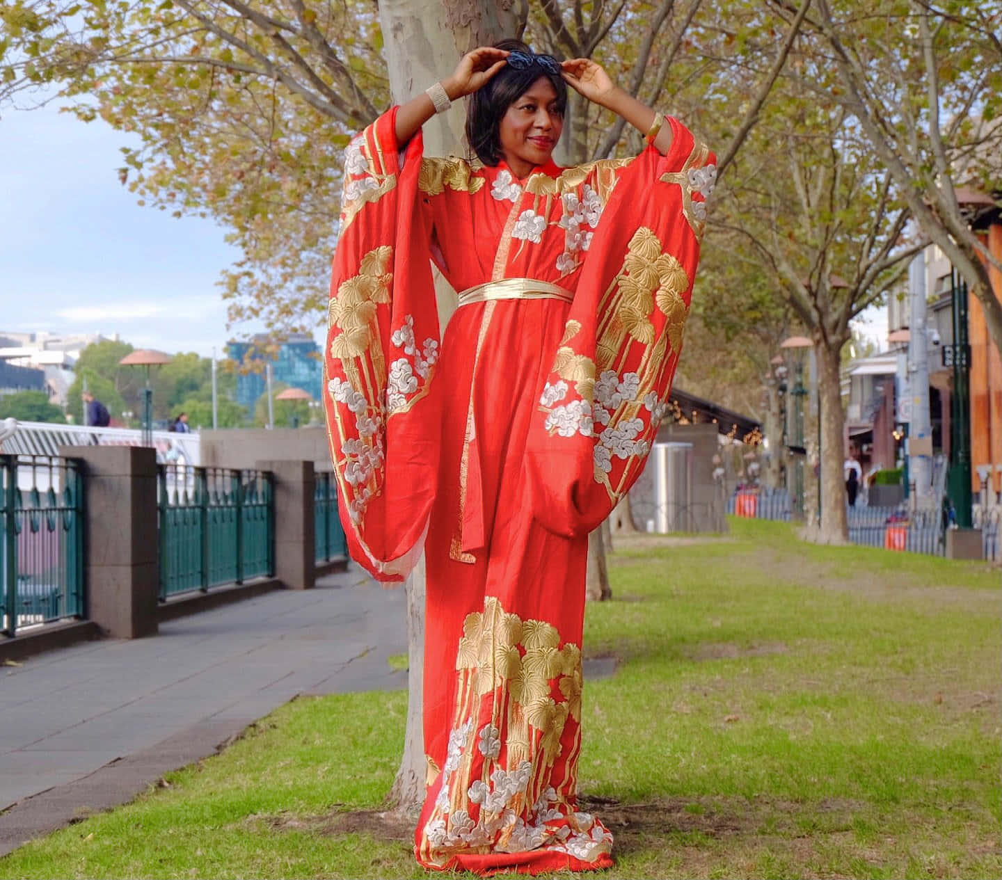 Kimono 桌布 壁紙 — 一位身穿鮮紅色和服、飾有金色和白色花卉刺繡的女性，在陽光明媚的城市公園中，優雅地站在枝葉繁茂的樹下。