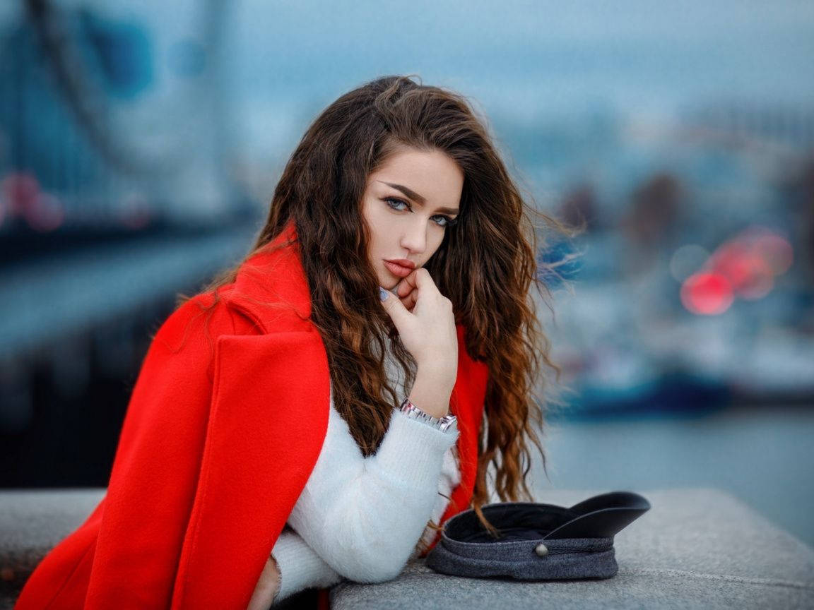 attitude 4K 桌布 壁紙 — 一位身穿鮮紅色外套的女性，若有所思地坐在水邊，身後是模糊的城市天際線。