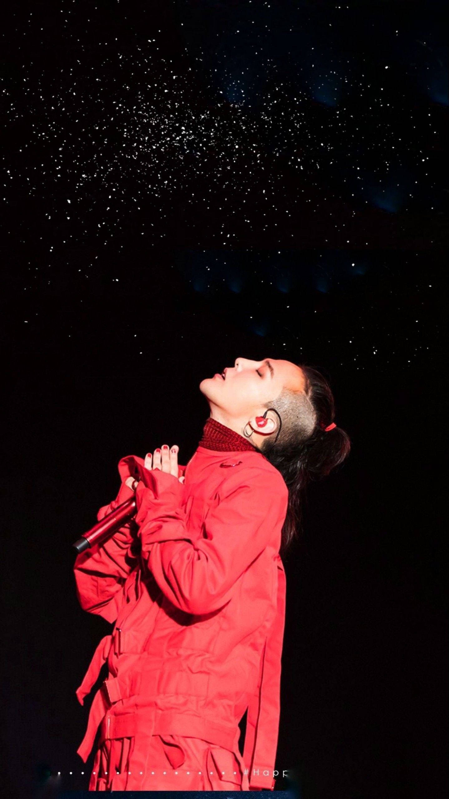 G-dragon 桌布 壁紙 — 一位身穿鮮紅色服裝的表演者雙手合十仰望，閃爍的粒子在黑暗的星空背景下飄落。