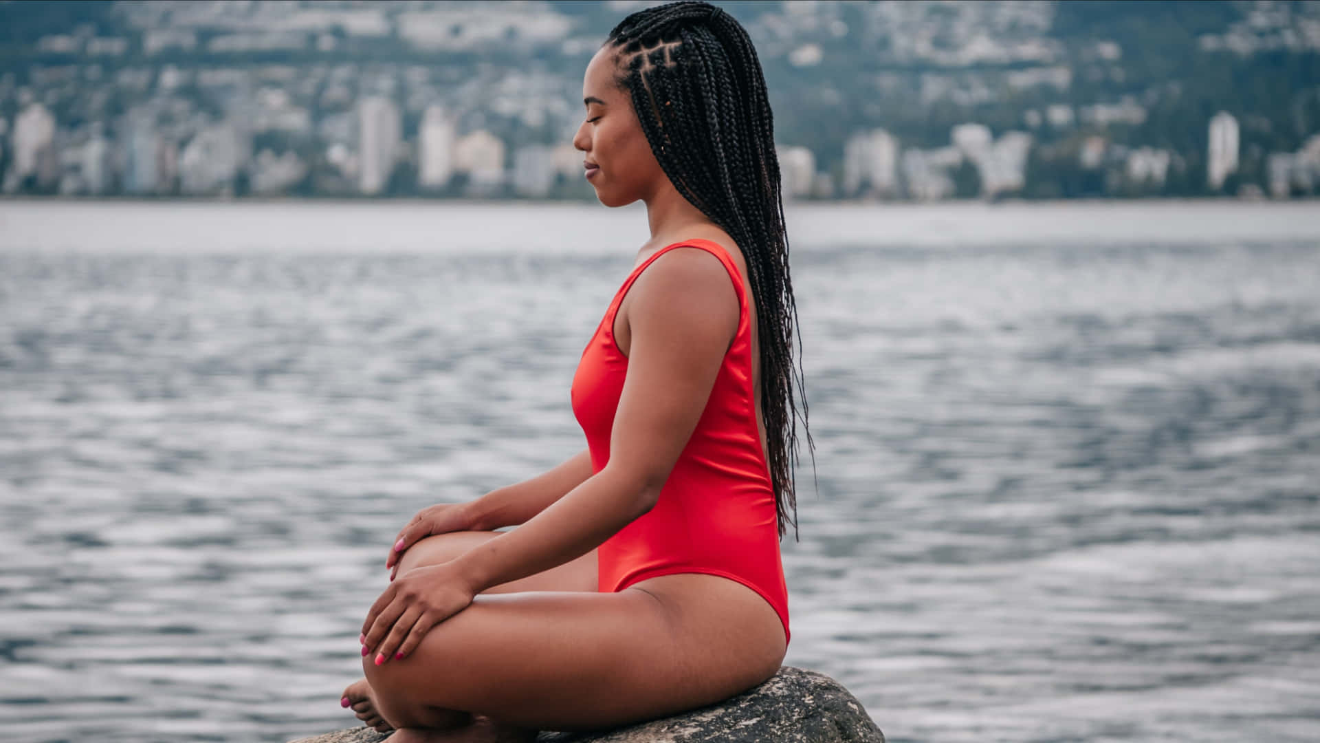 Meditation 桌布 壁紙 — 一位身穿鮮紅色泳衣的女性在水邊的岩石上平靜地冥想，背景是城市天際線。