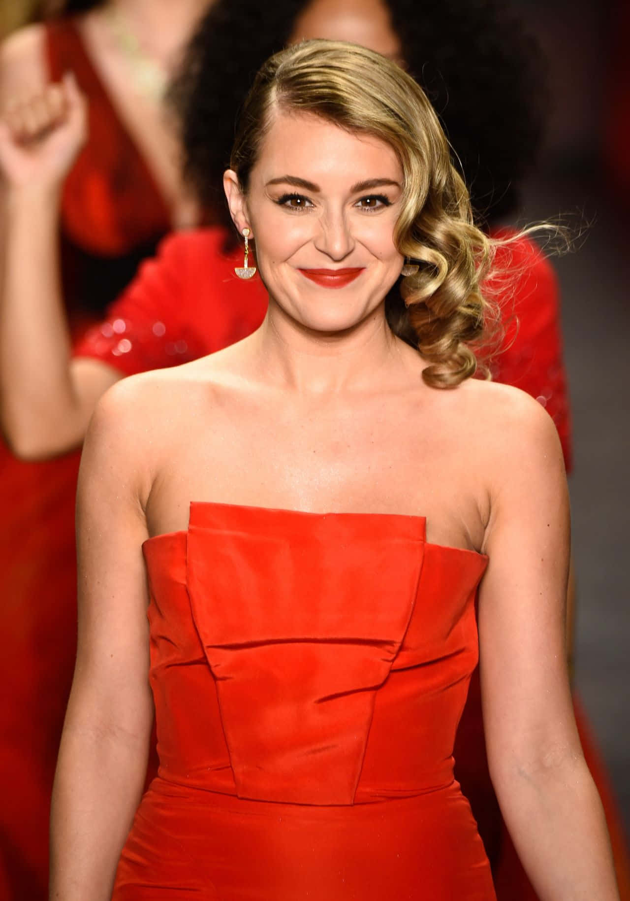 Alexa Penavega 桌布 壁紙 — 一位身穿鮮紅色無肩帶禮服的女士在時裝伸展臺上自信地微笑，身後有模糊的紅色模特兒。