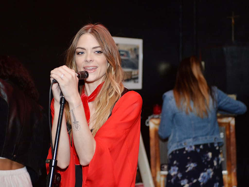 Jaime King 桌布 壁紙 — 一位身穿鮮紅色襯衫的女性在燈光昏暗的舞台上對著麥克風唱歌，背景有樂隊成員。
