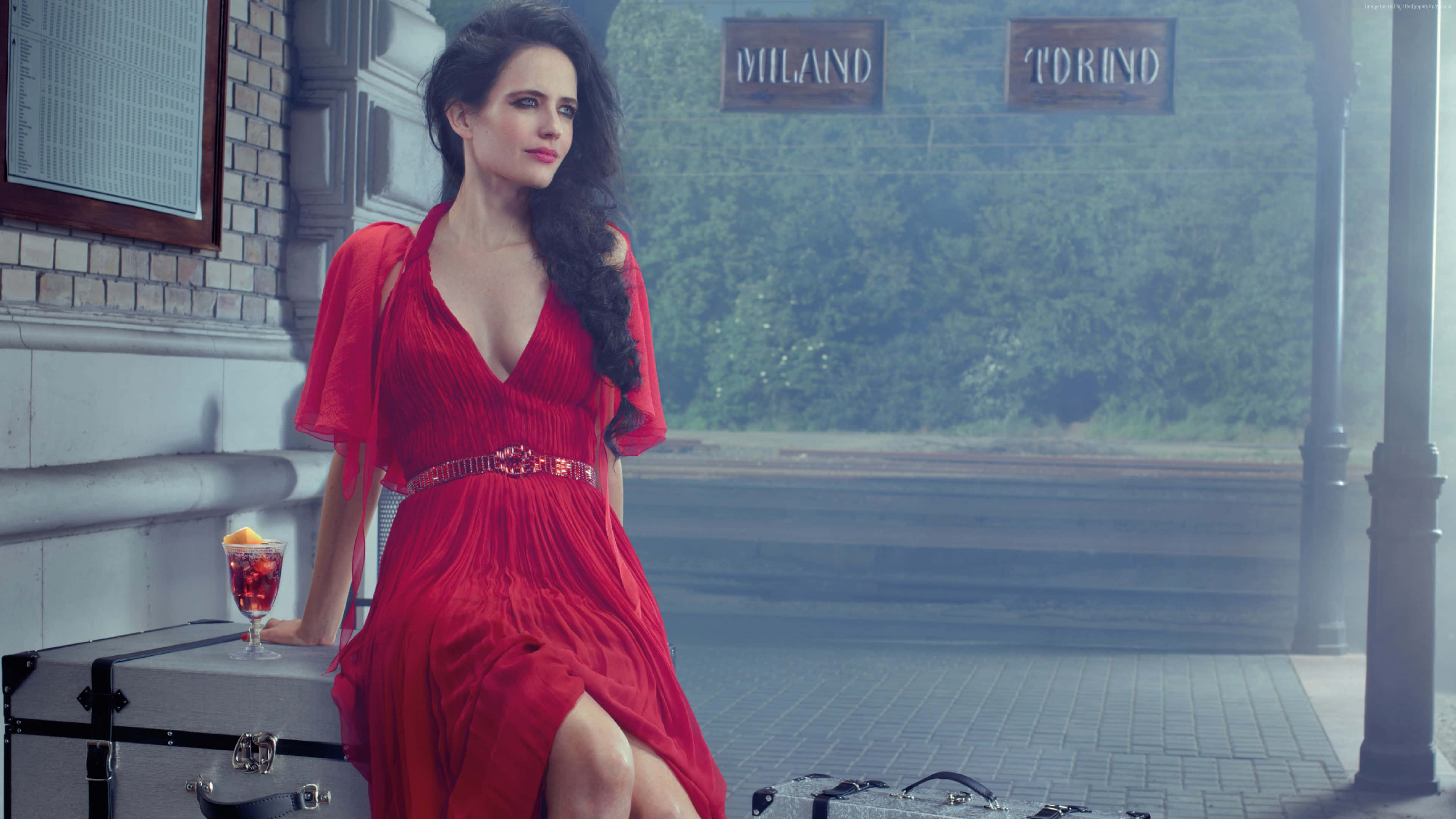 eva green 桌布 壁紙 — 一位身穿鮮紅色連衣裙的女性，優雅地坐在霧氣瀰漫的火車站月台上的行李箱上，旁邊有復古標誌。