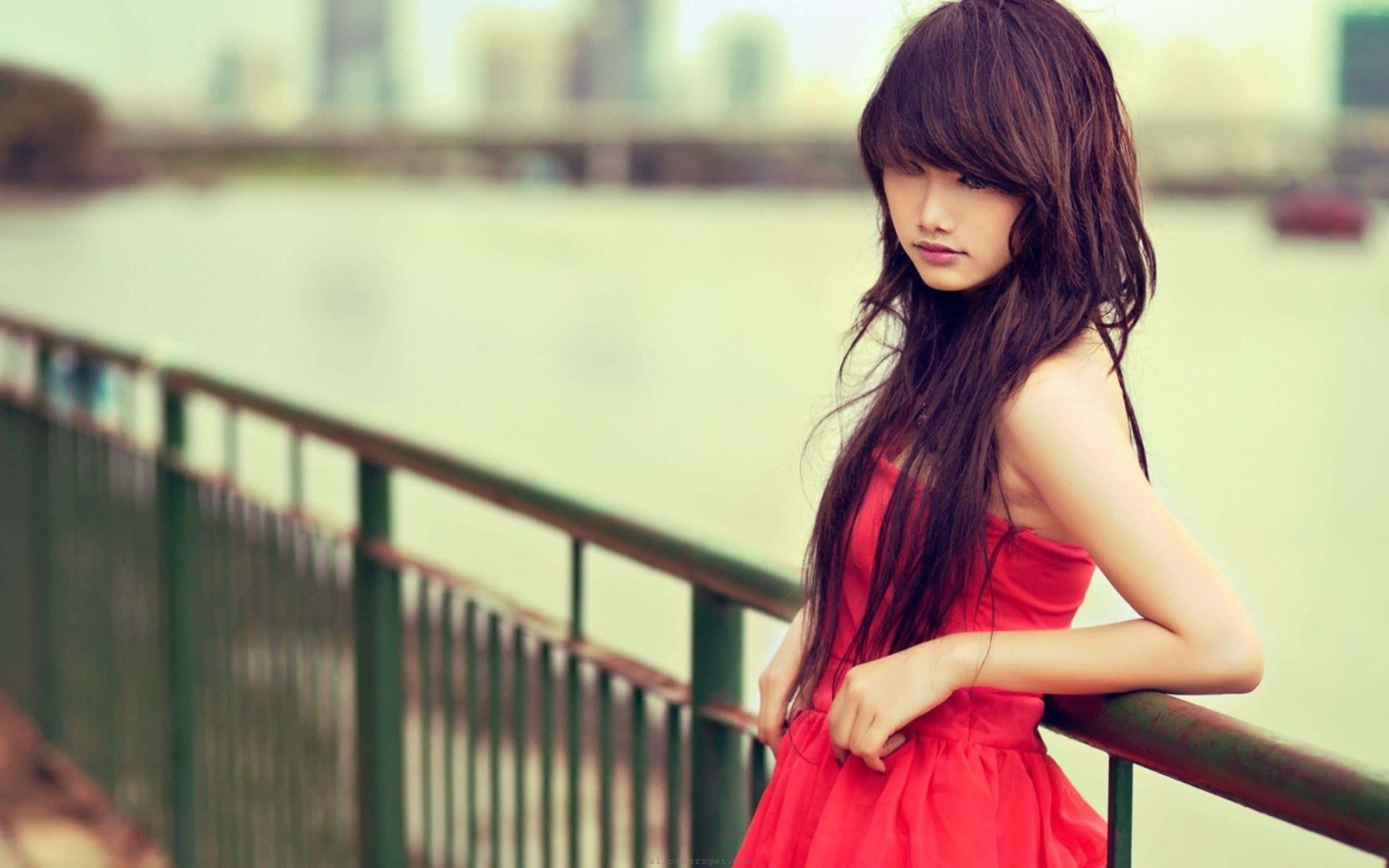 Beautiful Girls 桌布 壁紙 — 一位身穿鮮紅色連衣裙的年輕女子，若有所思地靠在平靜河流旁的綠色欄杆上，背景是模糊的城市天際線。