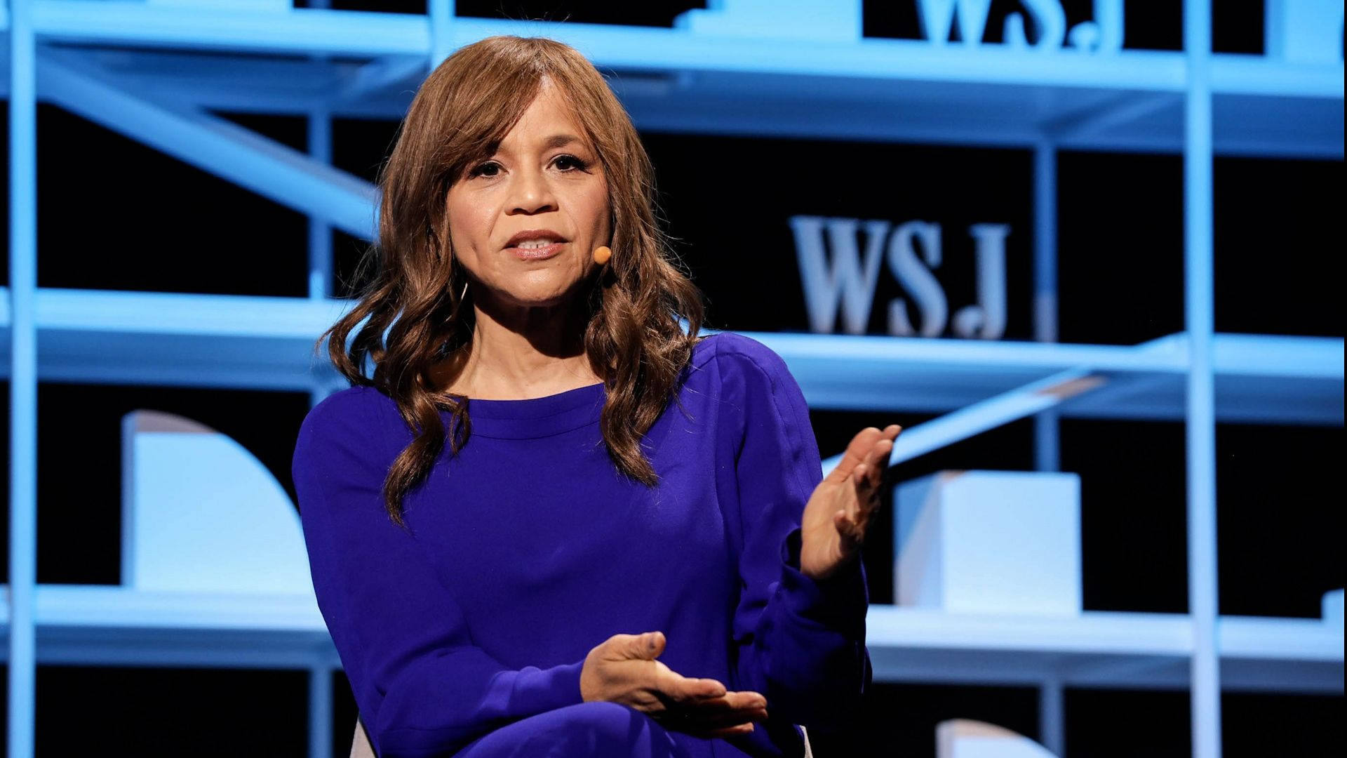 Celebrity 桌布 壁紙 — 一位身穿鮮紫色上衣的女性在背後帶有「WSJ」品牌的現代藍色燈光舞台上熱情演講。