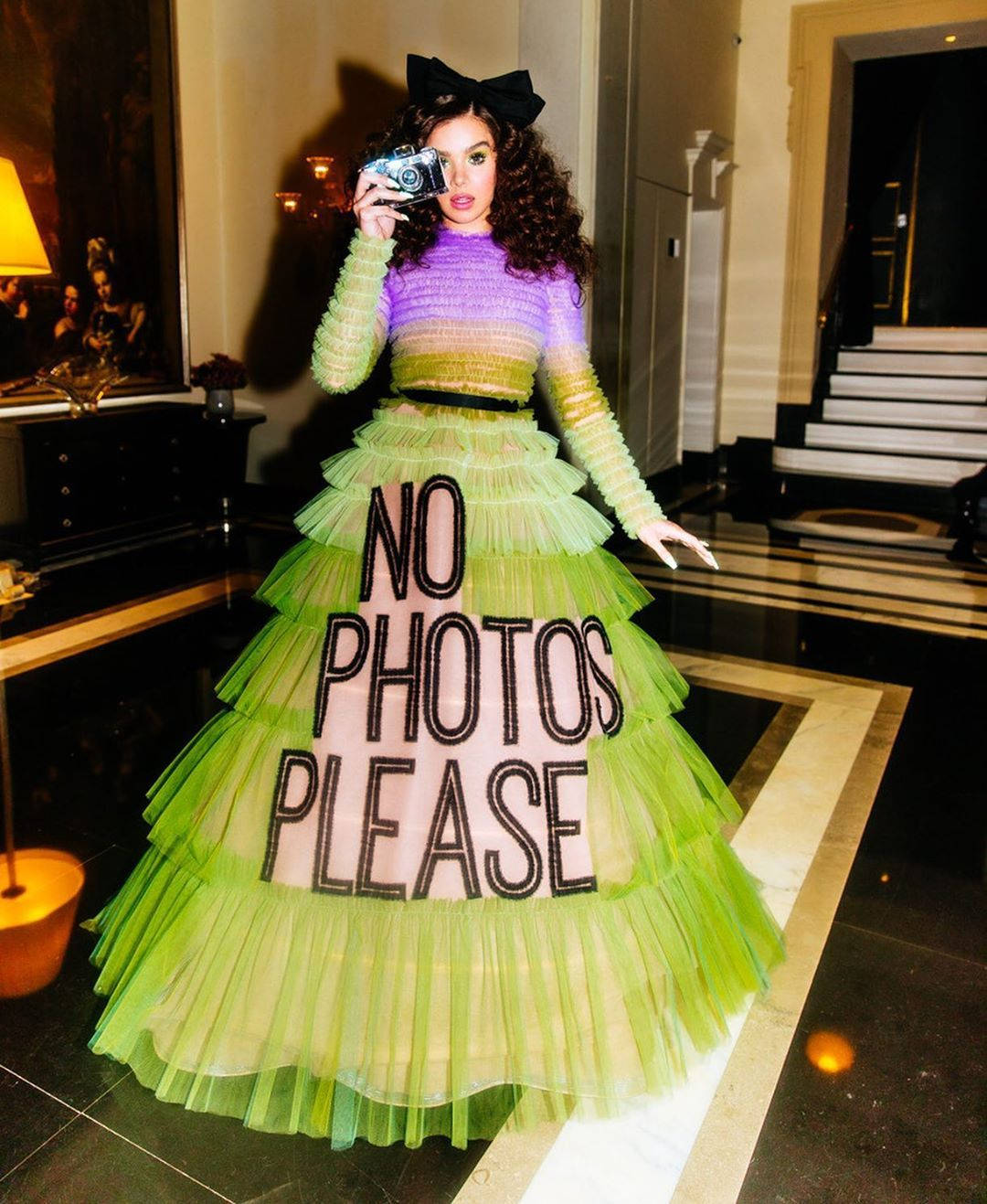 Hailee Steinfeld 桌布 壁紙 — 一位身穿鮮綠色和紫色分層長裙、帶有「NO PHOTOS PLEASE」字樣的女士，在豪華飯店大廳手持相機。