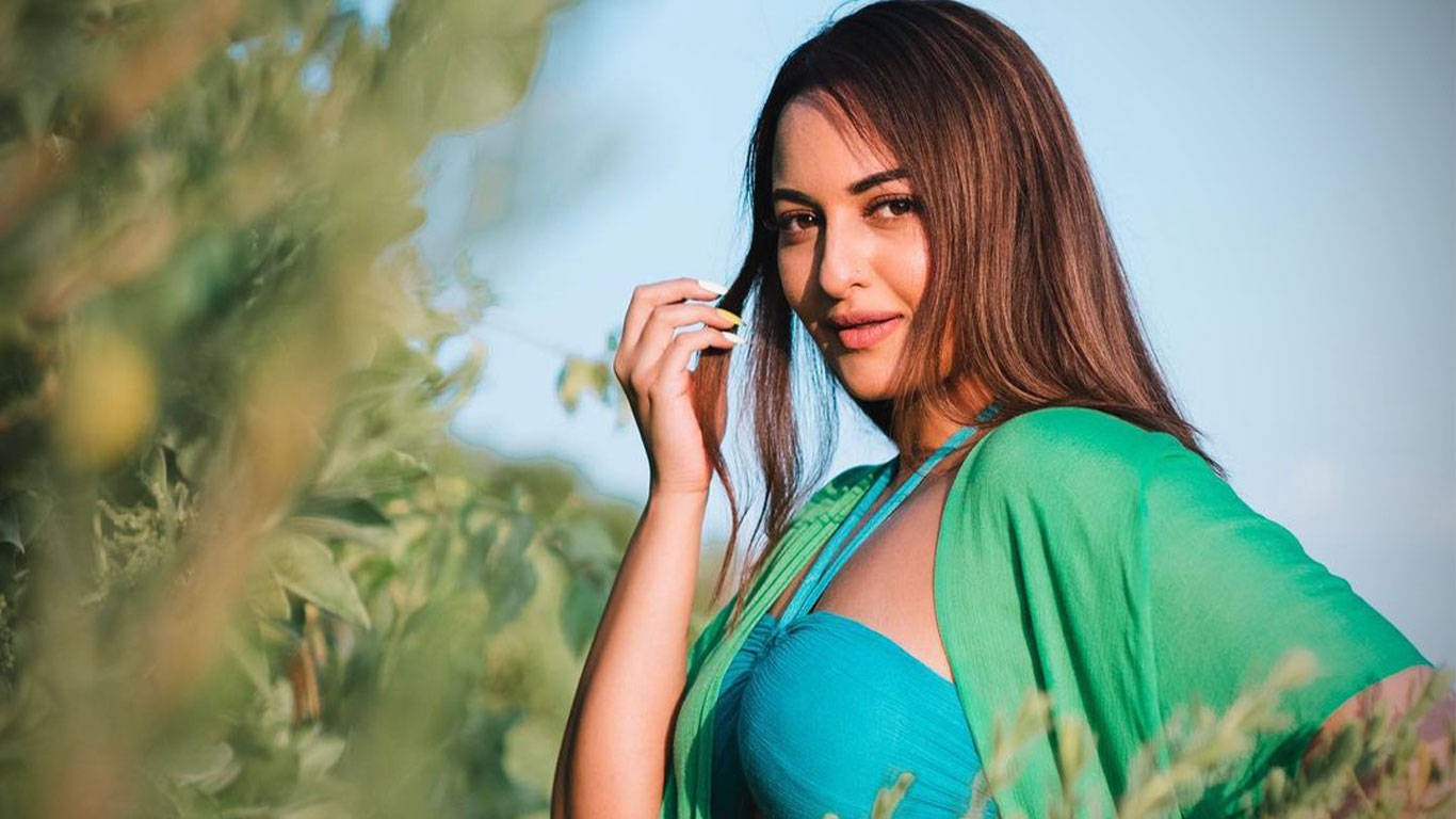 Sonakshi Sinha 桌布 壁紙 — 一位身穿鮮綠色和藍綠色服裝的女性，在明亮的日光下站在茂密的樹葉中，輕柔地觸摸頭髮，表情平靜。
