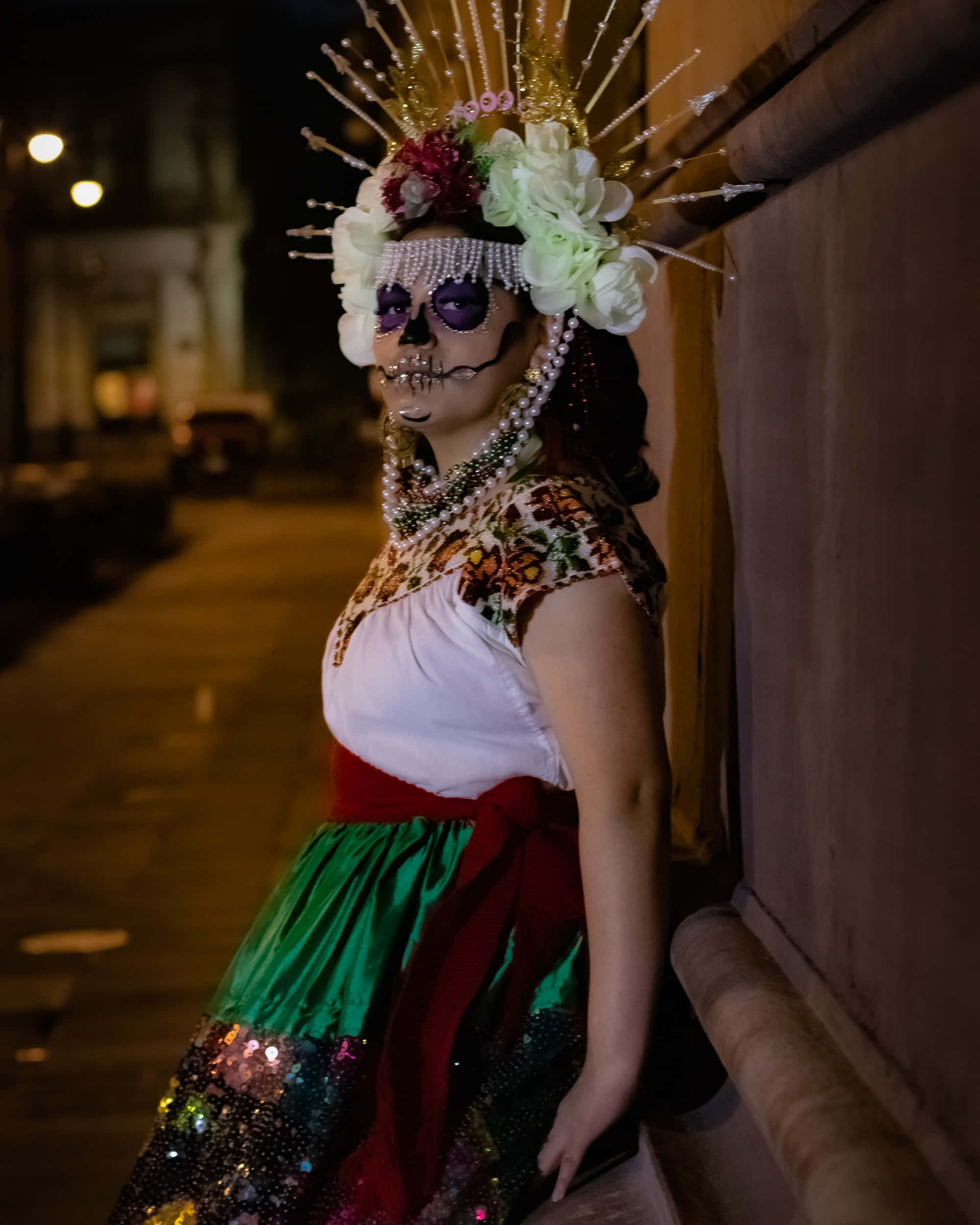Day Of The Dead 桌布 壁紙 — 一位身穿鮮豔亡靈節服飾、頭戴花卉頭飾、化著骷髏妝容的女性，在夜晚昏暗的城市小巷中站立。