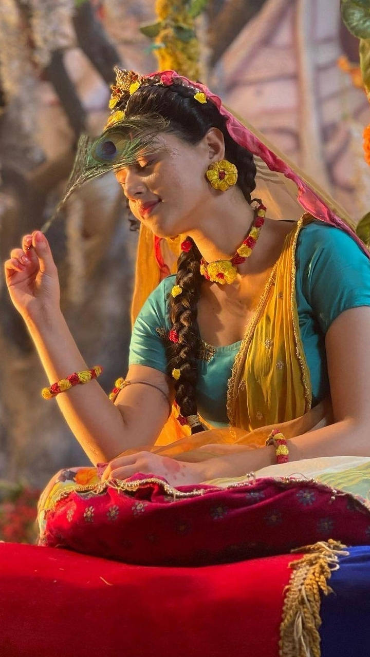 Mallika 桌布 壁紙 — 一位身穿鮮豔傳統印度服飾、飾有花朵和孔雀羽毛的女性，在溫暖的金色光芒中優雅地坐著。
