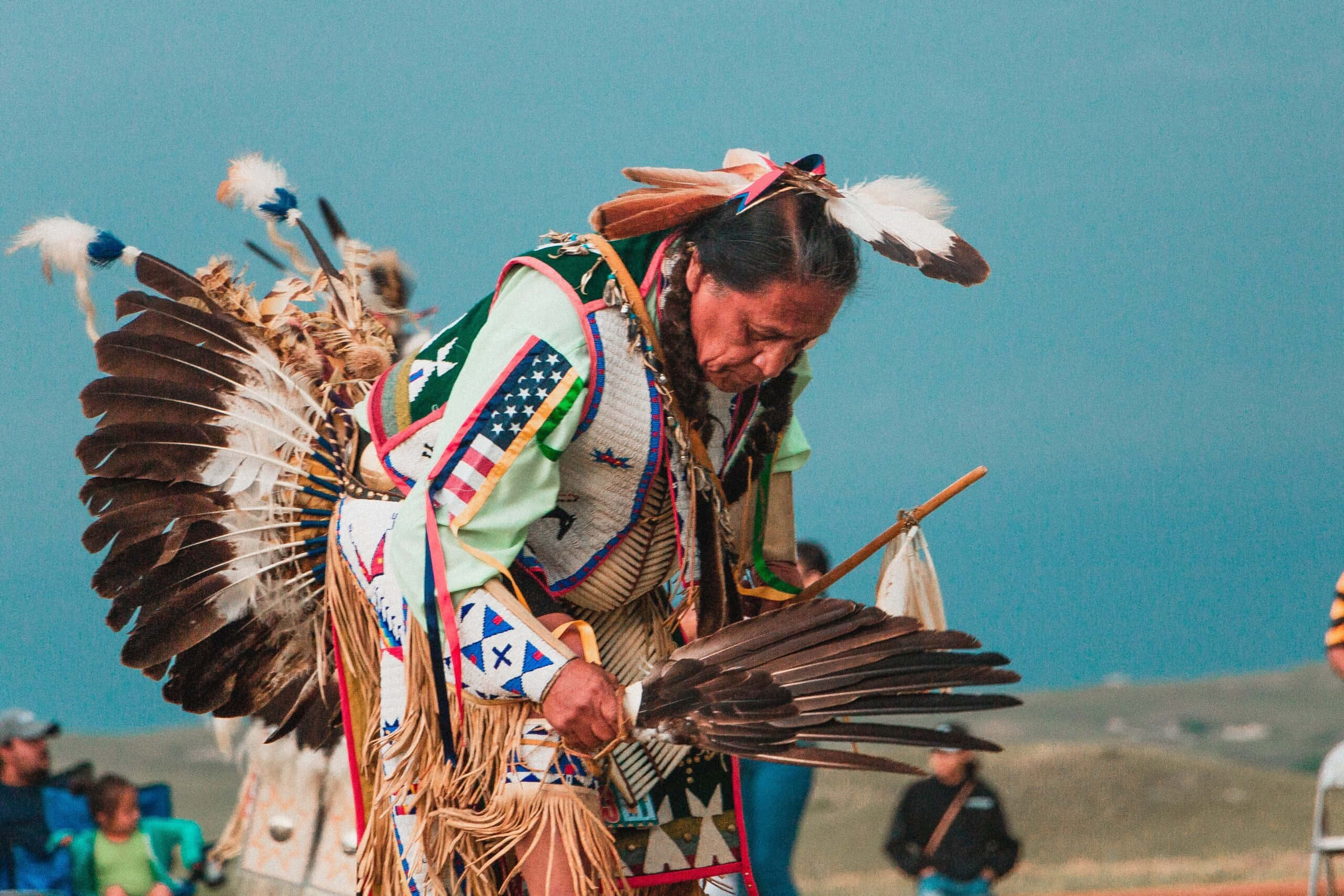 Native American 桌布 壁紙 — 一位身穿鮮豔傳統服飾、頭戴鷹羽頭飾的美洲原住民舞者，在晴朗的藍天下於戶外表演儀式舞蹈。