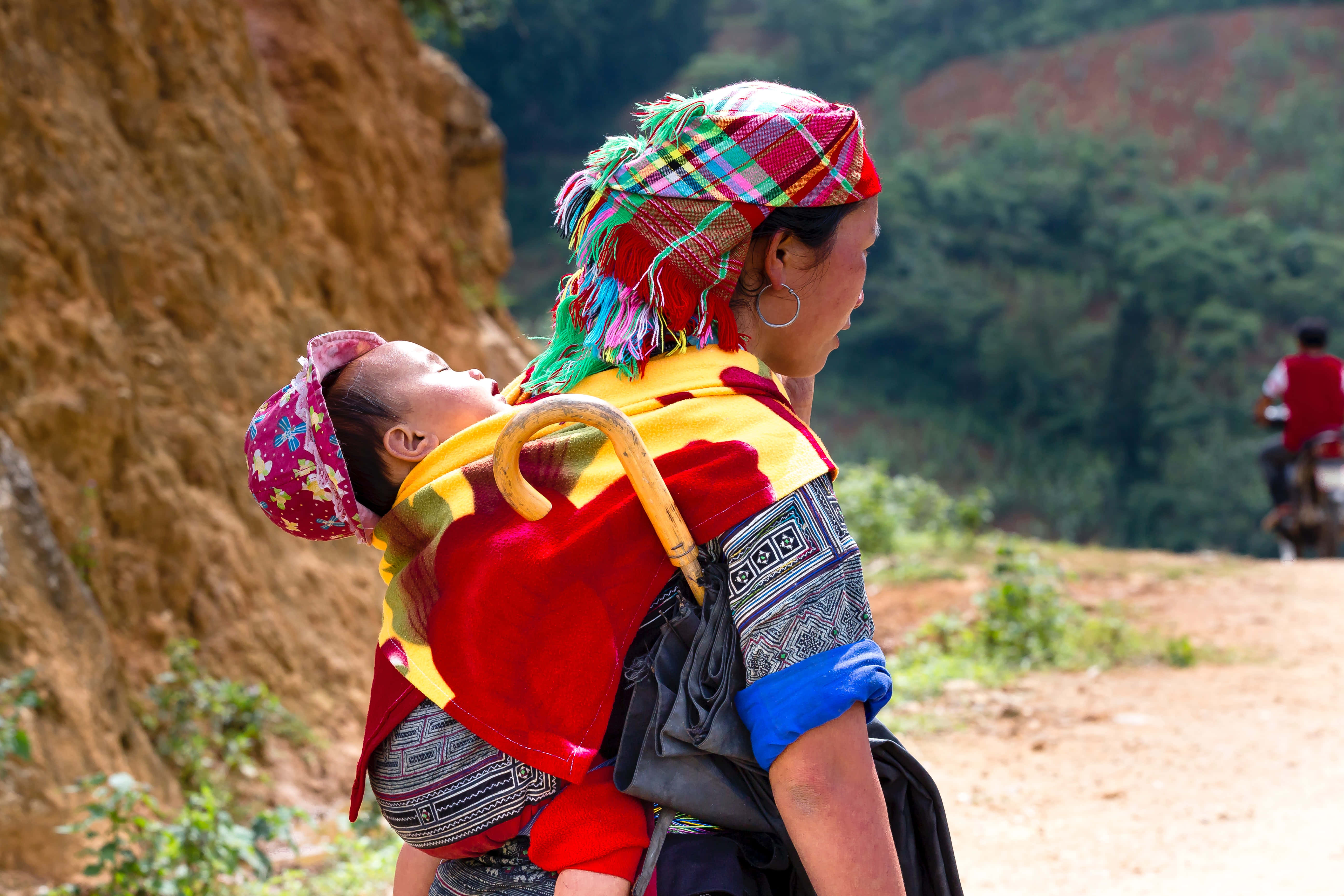Mom 桌布 壁紙 — 一位身穿鮮豔傳統服飾的女性，背著熟睡的嬰兒，沿著一條泥濘小路穿過崎嶇的山區景觀。