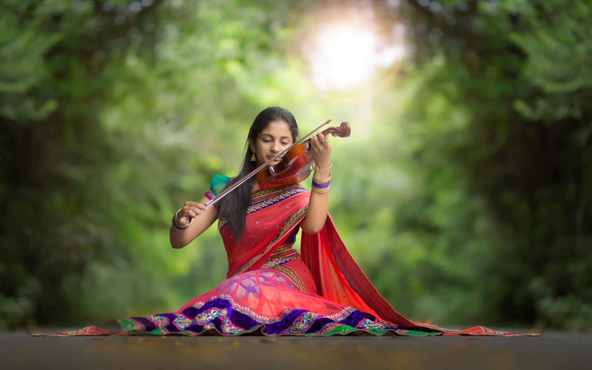 Indian 桌布 壁紙 — 一位身穿鮮豔傳統服飾的女性在柔和陽光穿過茂密綠樹的照射下演奏小提琴。