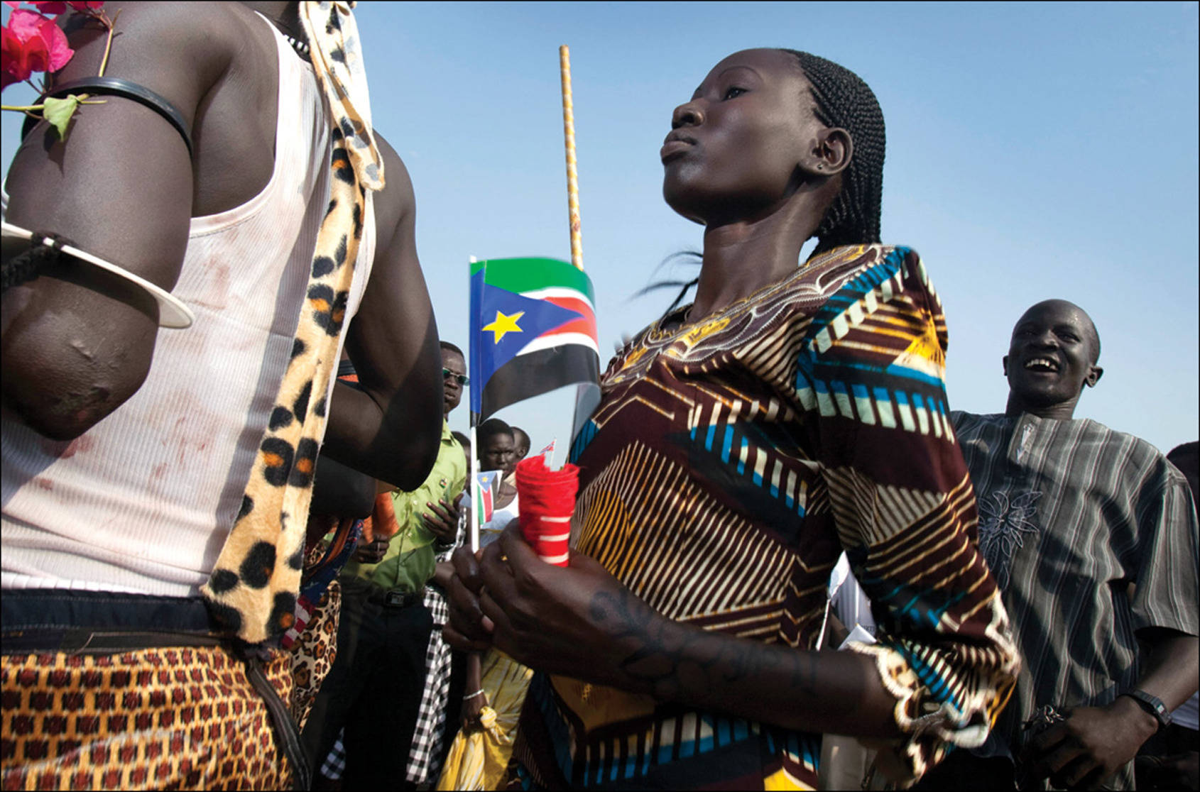 South Sudan 桌布 壁紙 — 一位身穿鮮豔圖案服裝的女性，在陽光明媚的人群中手持南蘇丹國旗，參加慶祝活動。