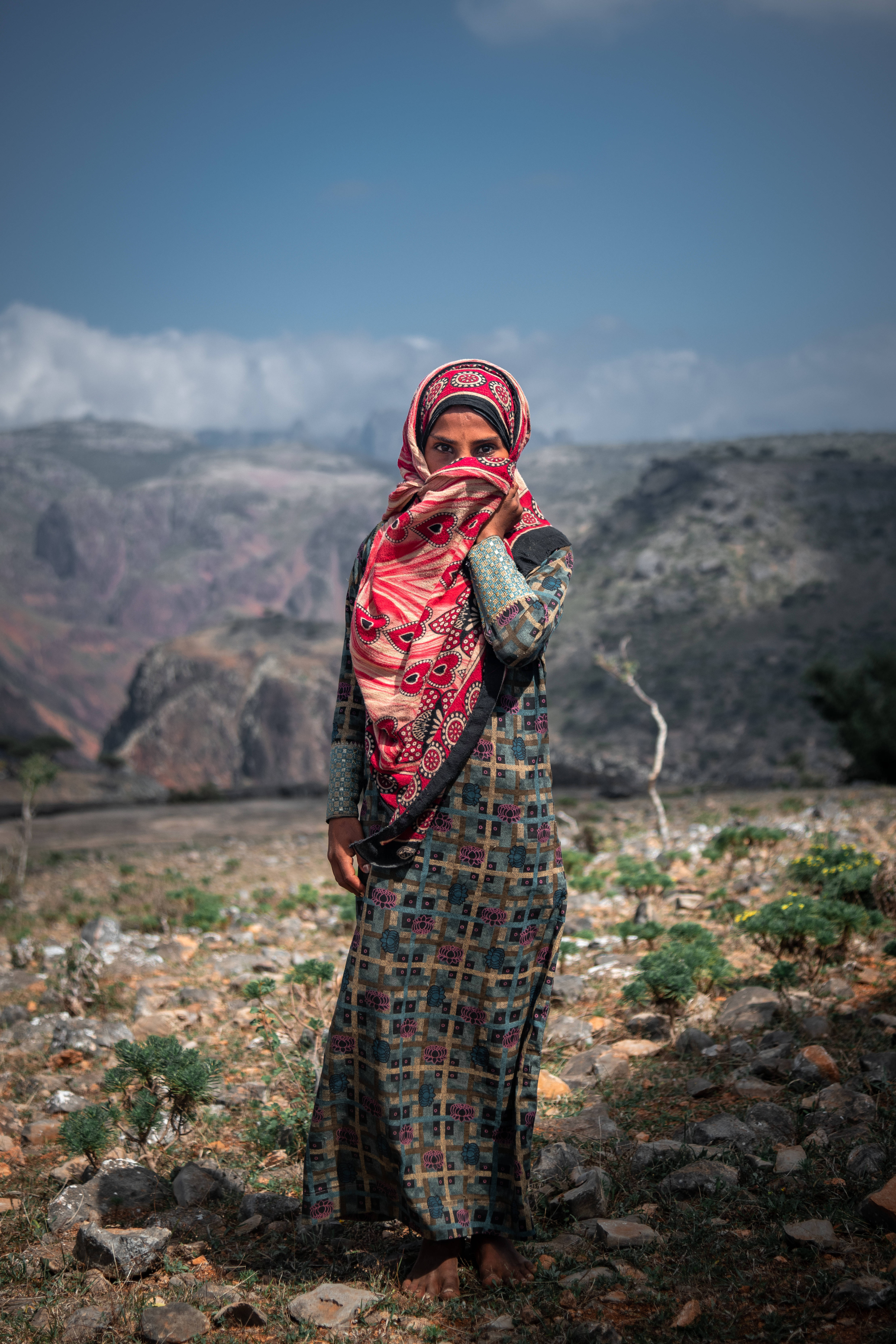 Yemen 桌布 壁紙 — 一位身穿鮮豔圖案服飾的女性，在晴朗的藍天下赤腳站在岩石山區地形中。