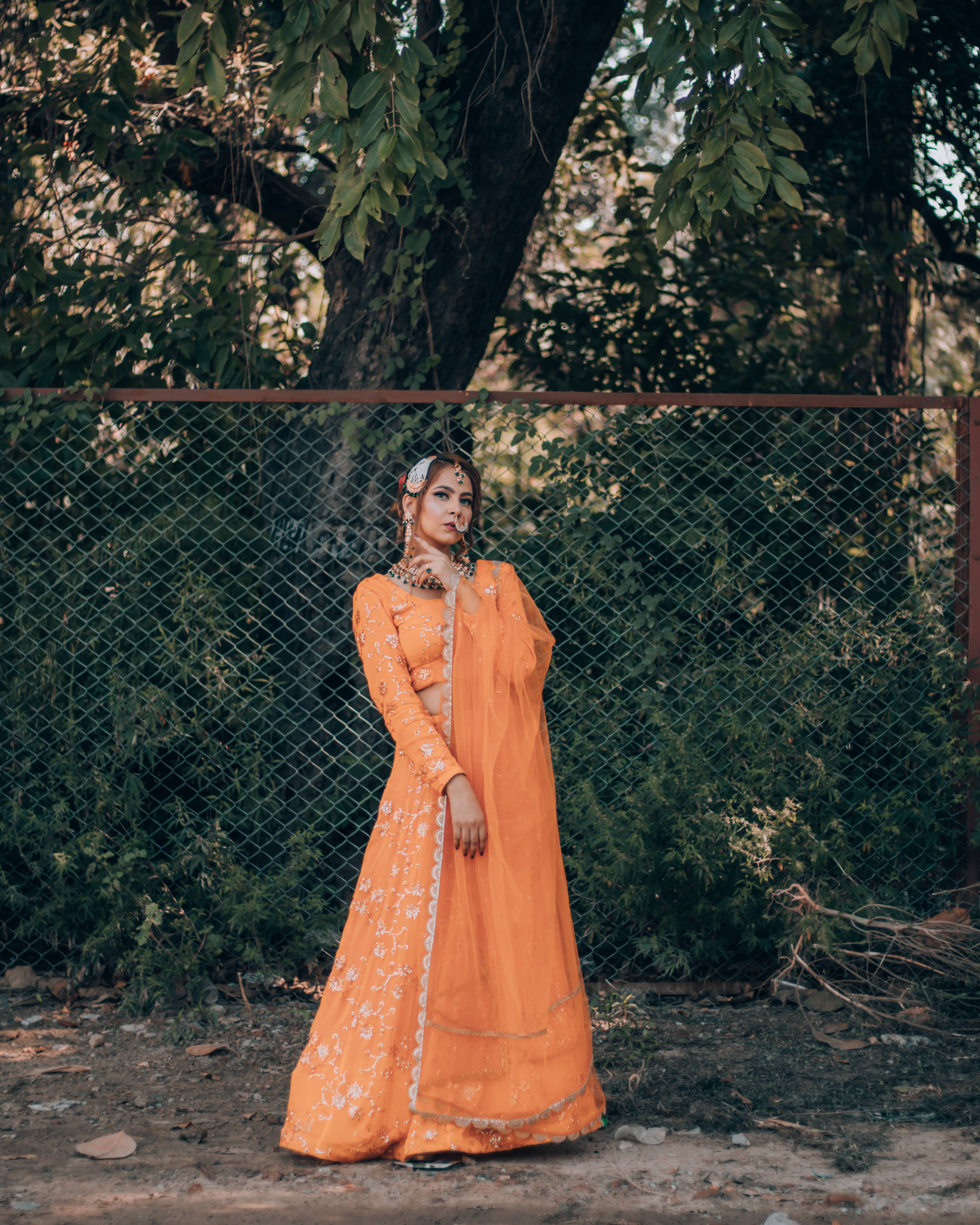 Indian 桌布 壁紙 — 一位身穿鮮豔橙色刺繡 lehenga 的女性，在樹葉茂密的樹木過濾下的斑駁陽光中，優雅地站在鐵絲網圍欄旁。