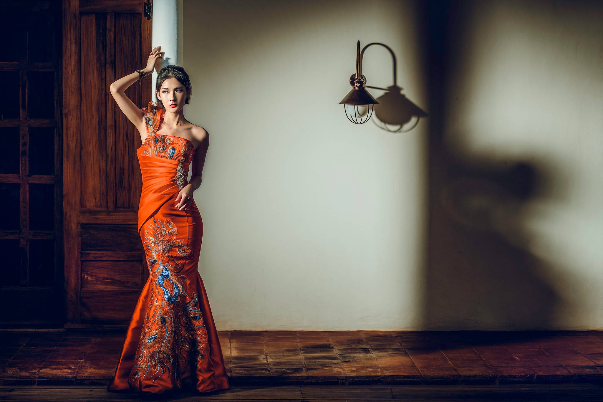 Dress 桌布 壁紙 — 一位身穿鮮豔橙色刺繡長裙的女性，優雅地站在一個光線溫暖、帶有木門和復古壁燈的質樸房間裡。