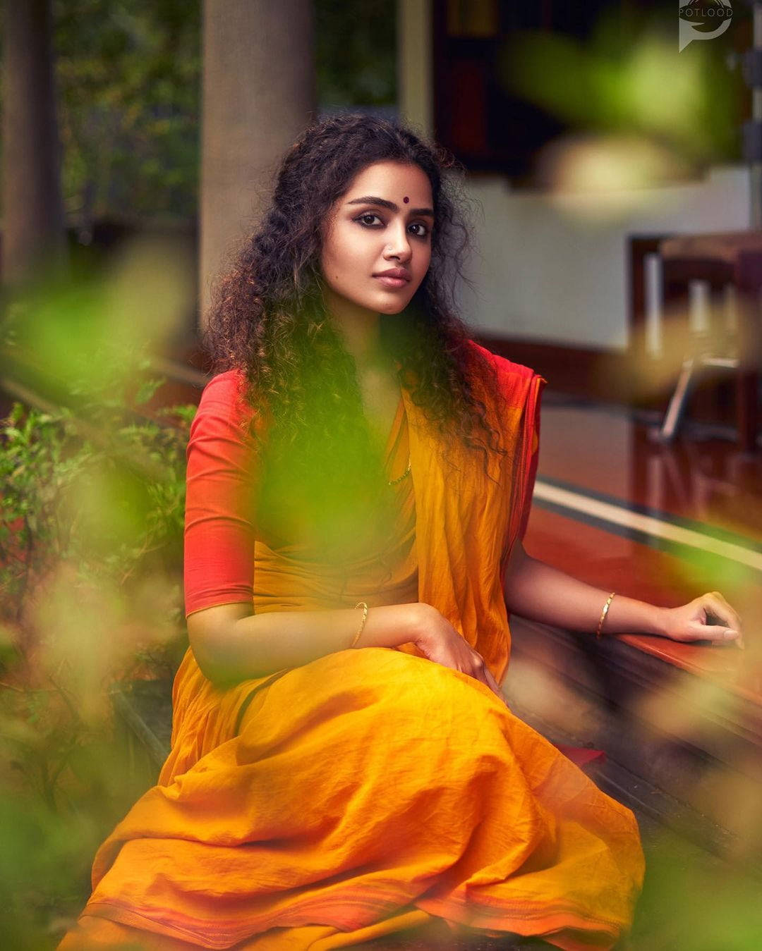 anupama parameswaran 桌布 壁紙 — 一位身穿鮮豔橙色和紅色紗麗的女性，優雅地坐在被模糊綠色樹葉環繞的木長椅上。