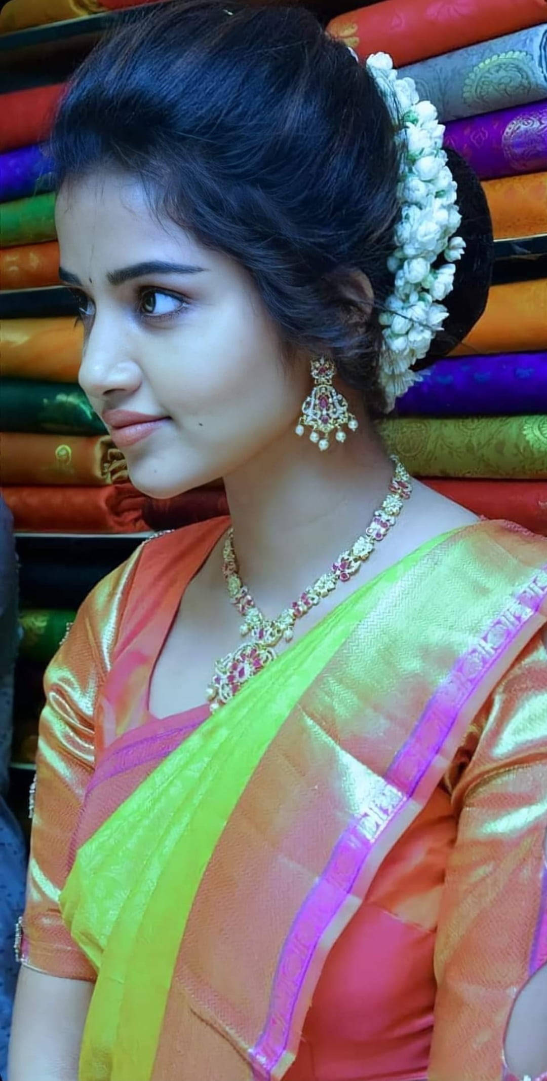 anupama parameswaran 桌布 壁紙 — 一位身穿鮮豔橙色和綠色紗麗、飾有白色花朵和黃金首飾的女性，在彩色布料卷旁若有所思地凝視。