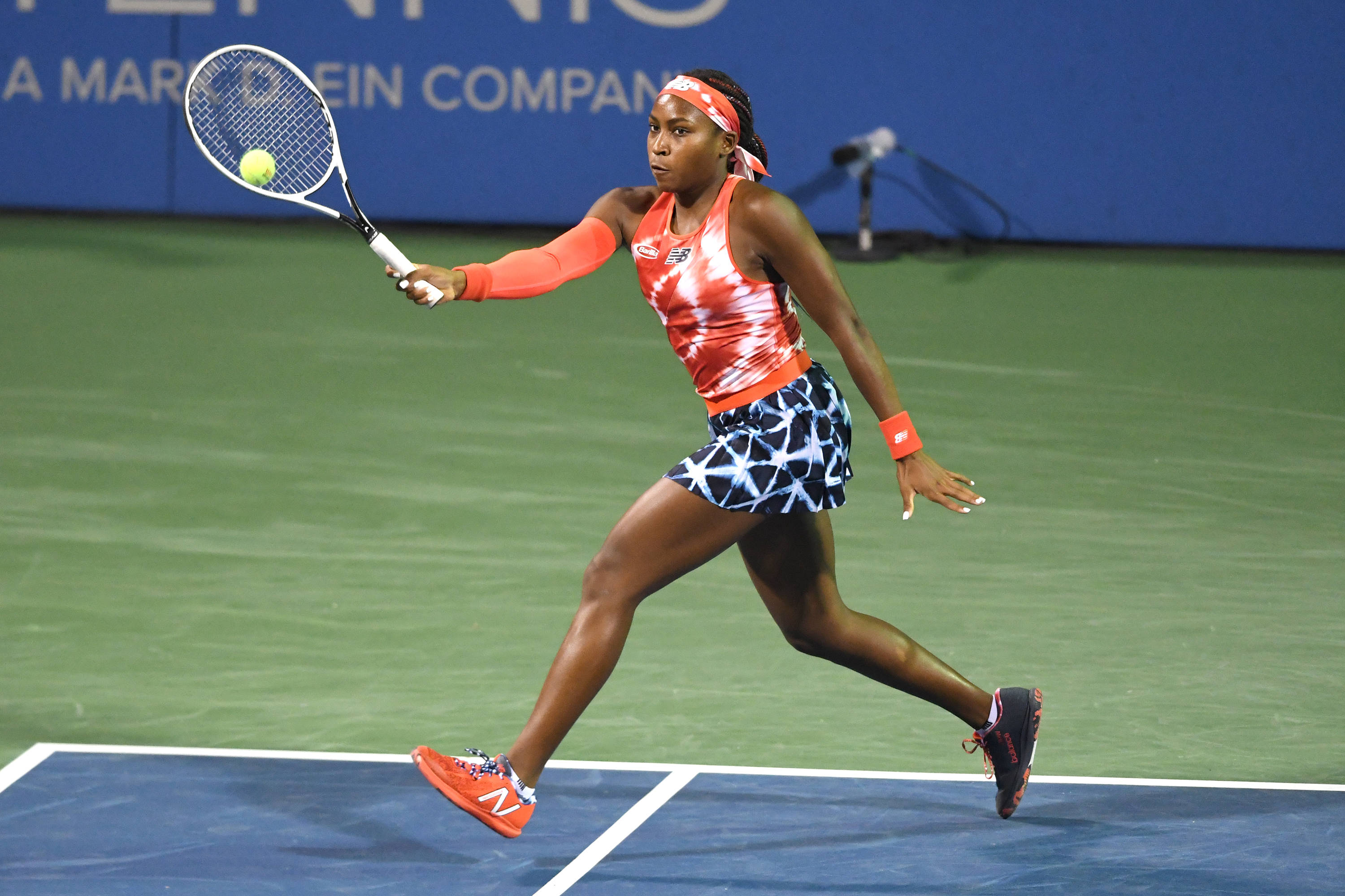 Coco Gauff 桌布 壁紙 — 一位身穿鮮豔橙色和藍色裝備的女網球選手在明亮燈光下的綠色球場上向前猛衝擊打黃色球。
