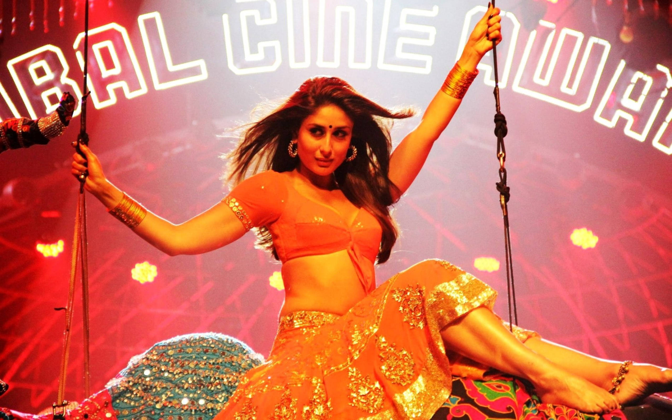 Kareena 桌布 壁紙 — 一位身穿鮮豔橙色和金色傳統服飾的女性，在紅色燈光下閃閃發光的舞台上充滿活力地表演。