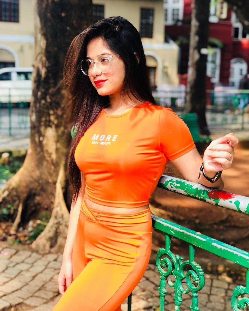Indian 桌布 壁紙 — 一位身穿鮮豔橙色短版上衣和緊身褲的女性，在陽光明媚的城市公園中倚靠著一條風化的綠色欄杆。