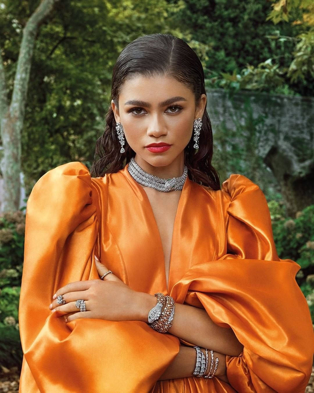 Zendaya 桌布 壁紙 — 一位身穿鮮豔橙色緞面禮服、佩戴閃亮鑽石珠寶的女性，自信地站在茂密的綠色森林背景下。