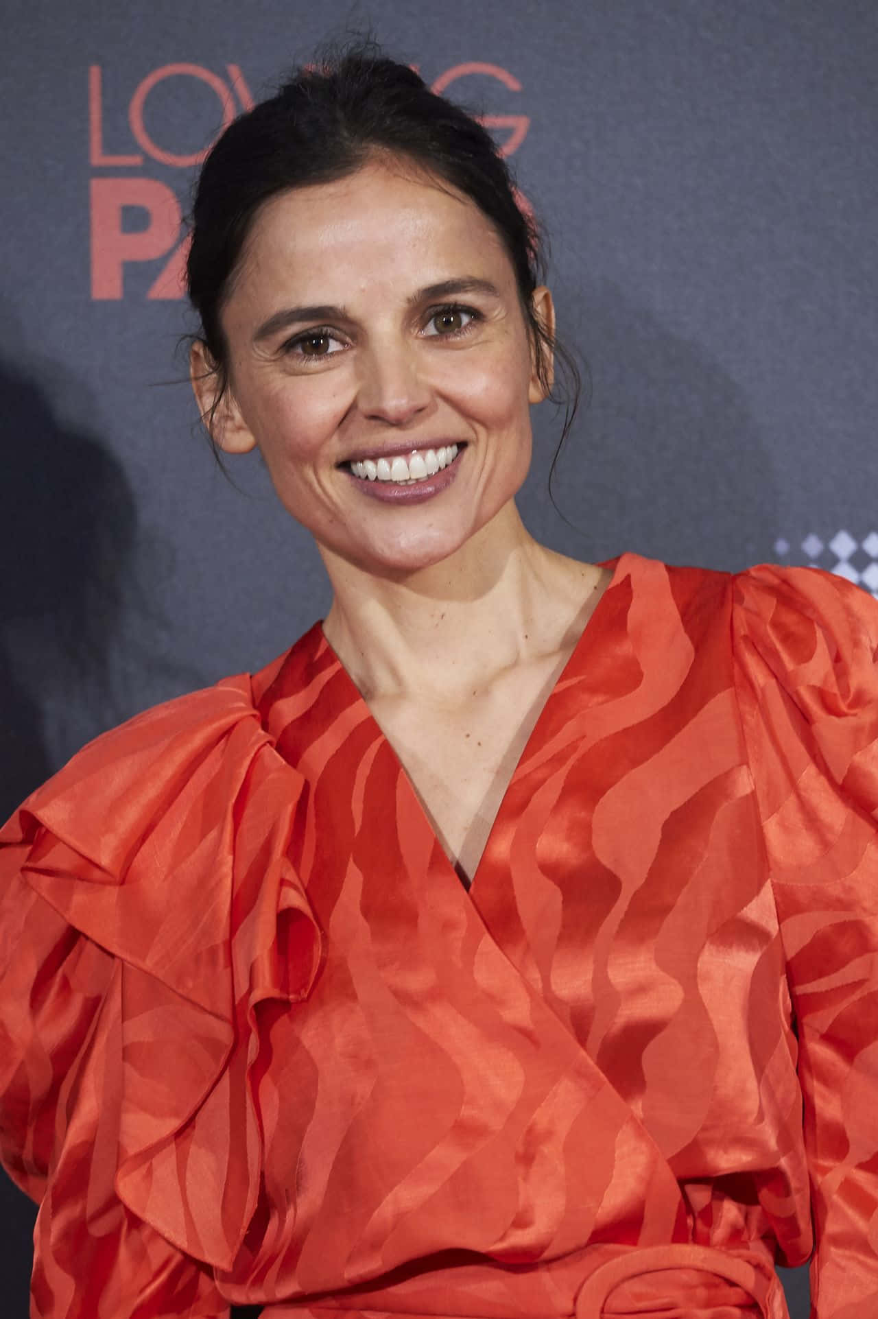 Elena Anaya 桌布 壁紙 — 一位身穿鮮豔橙色荷葉邊連衣裙的容光煥發的女性在黑暗背景的紅地毯活動中燦爛微笑。