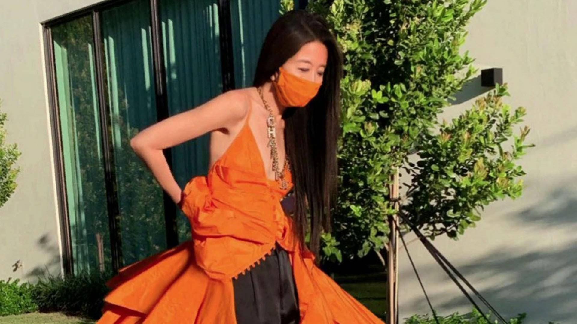 Asian Woman 桌布 壁紙 — 一位身穿鮮豔橙色連衣裙和相配口罩的女性在戶外自信地擺姿勢，旁邊有綠植和現代建築。