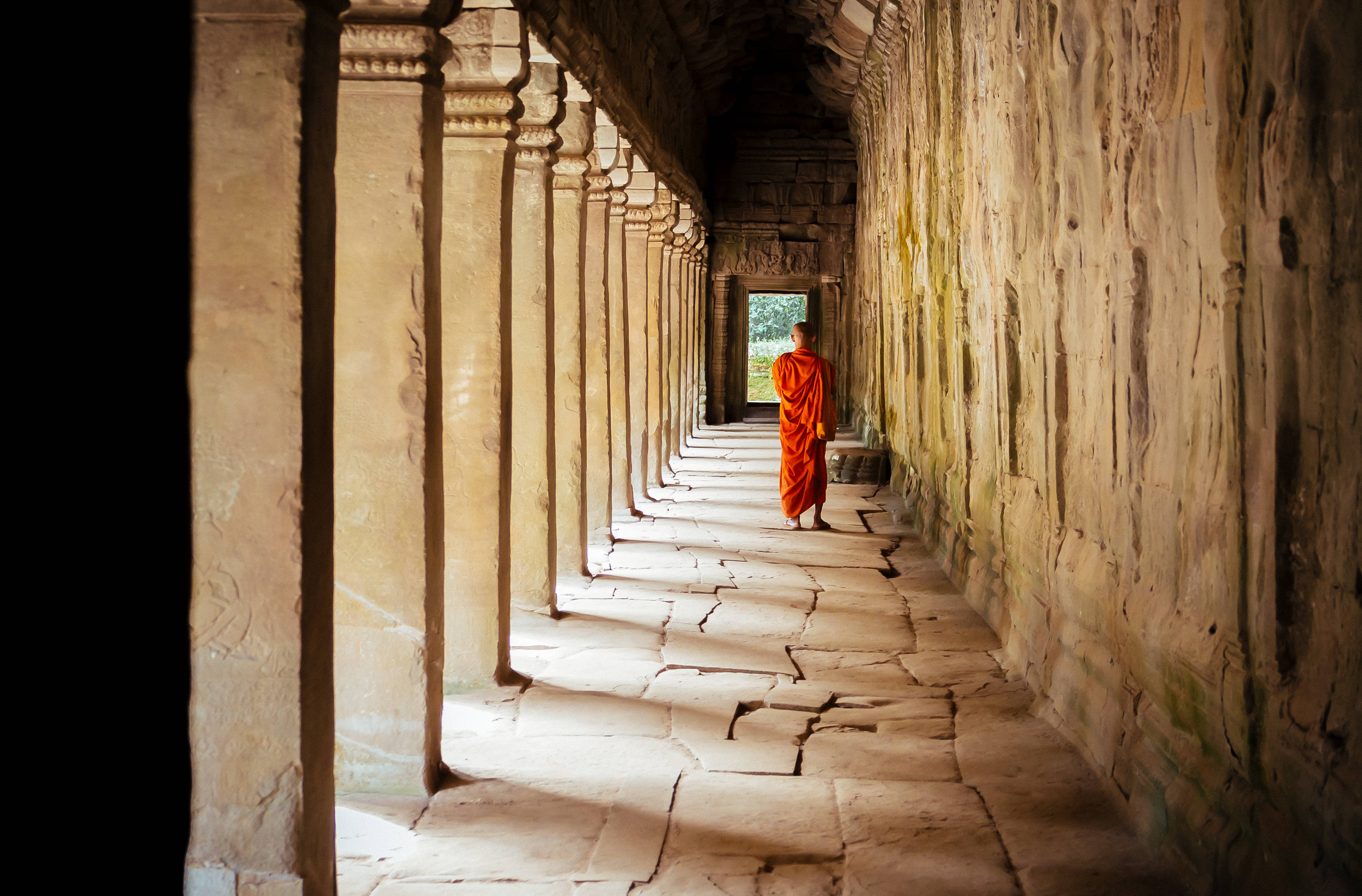 Cambodia 桌布 壁紙 — 一位身穿鮮豔橙色長袍的僧侶，沿著一條由古老柱子和斑駁陽光構成的長長石廟走廊行走。