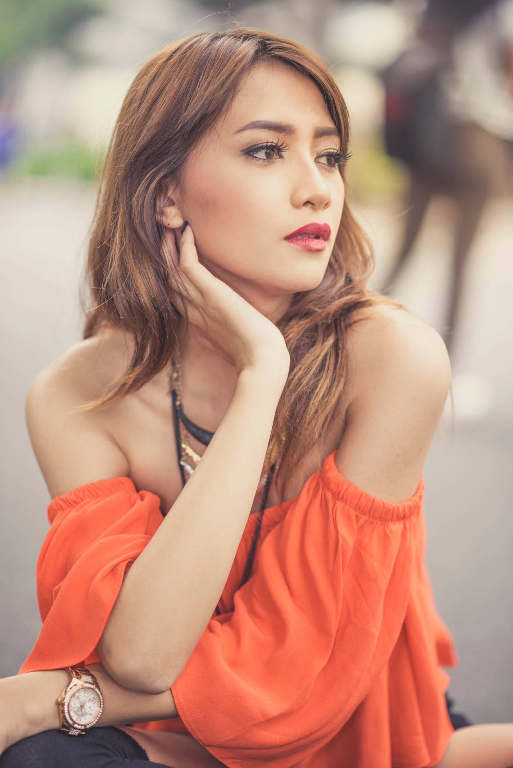 female 桌布 壁紙 — 一位身穿鮮豔橙色露肩上衣的女性，在柔和模糊的城市背景下，將手靠近臉龐，若有所思地擺姿勢。