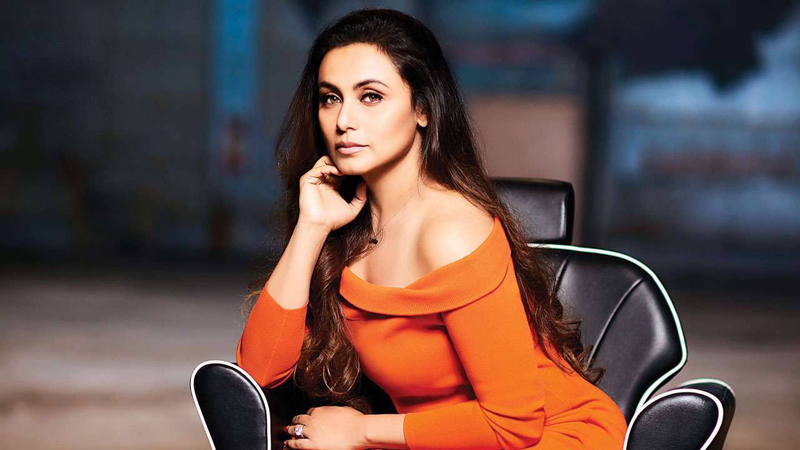 Rani Mukerji 桌布 壁紙 — 一位身穿鮮豔橙色露肩連衣裙的女性，端莊地坐在帶有白色飾邊的黑色椅子上，若有所思地凝視著鏡頭。