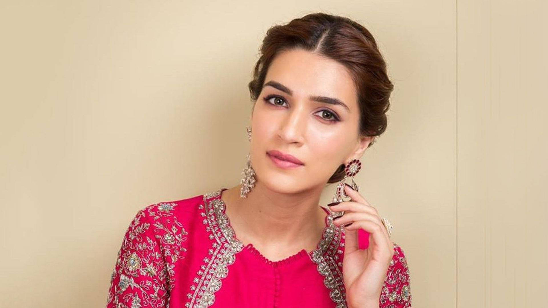 Kriti 桌布 壁紙 — 一位身穿鮮豔粉紅色刺繡服裝、帶有複雜銀色細節和大而華麗耳環的女性，在柔和的米色牆壁前優雅地擺姿勢。