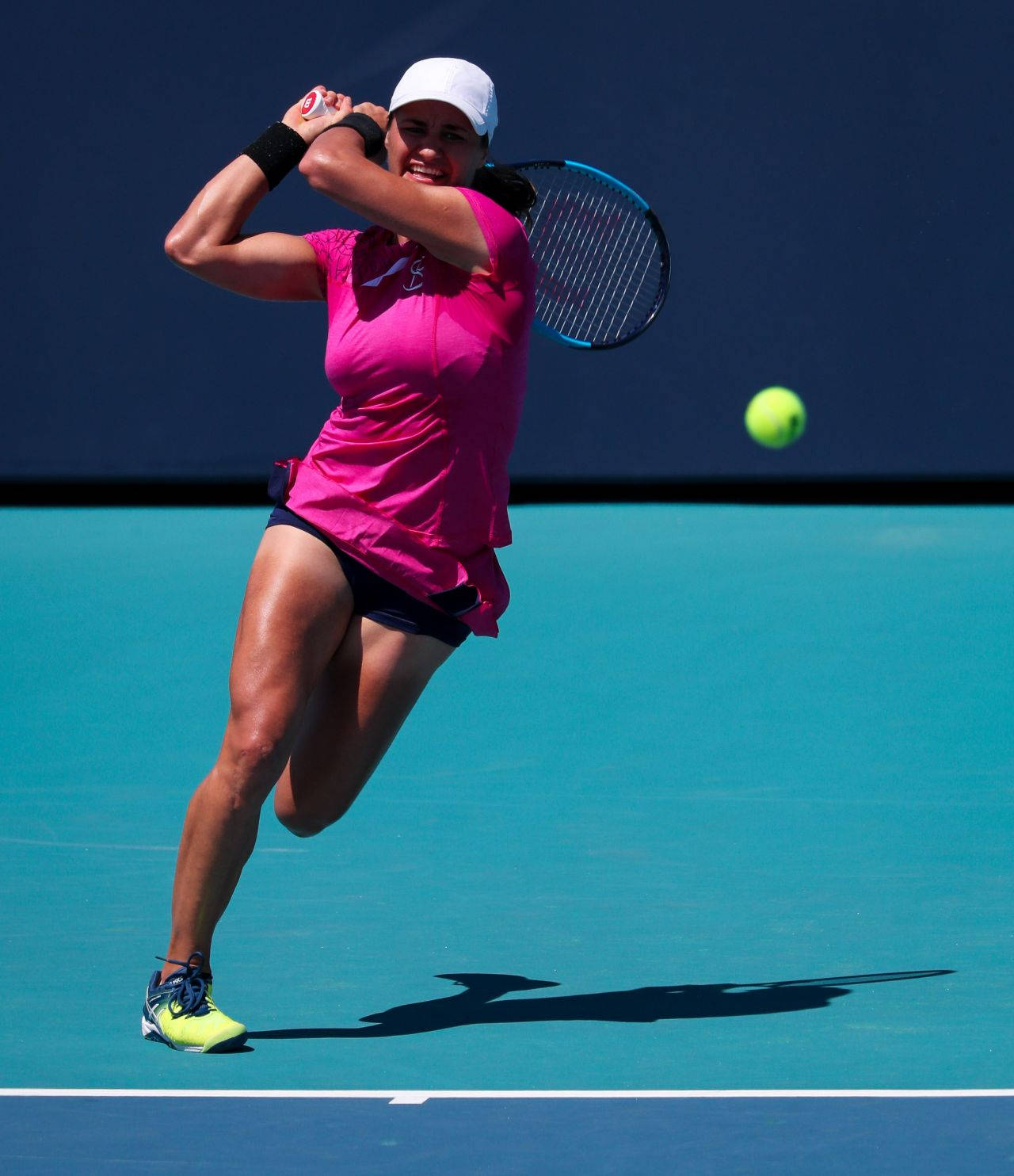 Tennis 桌布 壁紙 — 一位身穿鮮豔粉紅色服裝的女網球運動員，在晴朗的日光下，在藍綠色的球場上揮拍擊打一個亮黃色的球。