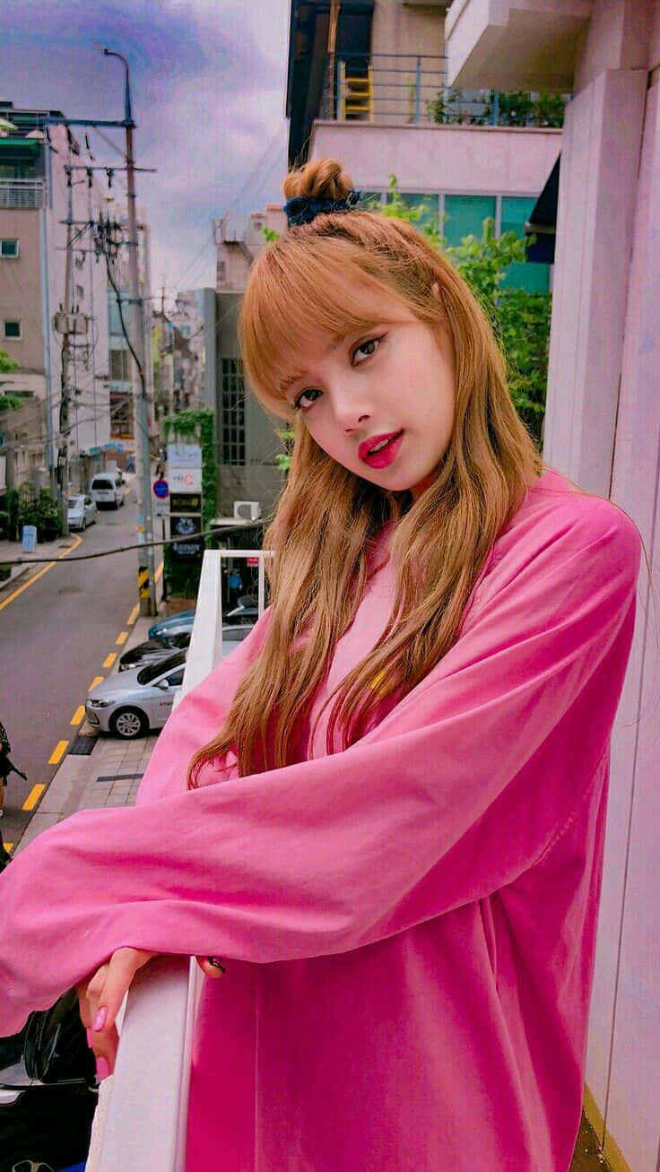 Lalisa 桌布 壁紙 — 一位身穿鮮豔粉紅色服裝的年輕女性，自信地在城市陽台上擺姿勢，身後是繁忙的街道。
