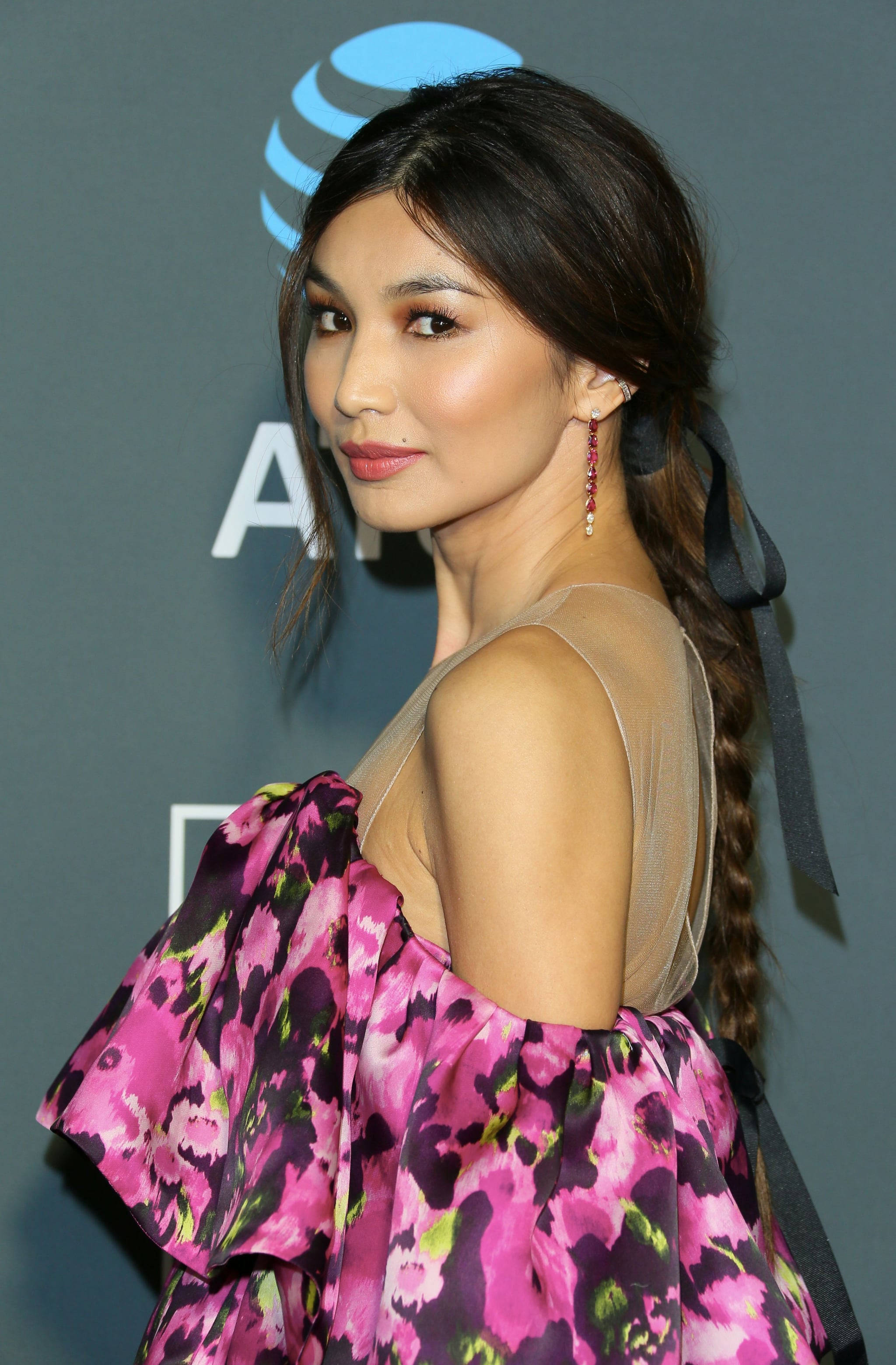 Gemma Chan 桌布 壁紙 — 一位身穿鮮豔粉紅色花卉連衣裙、側辮子和紅色耳環的女性在活動中對著帶有標誌的灰色背景擺姿勢。