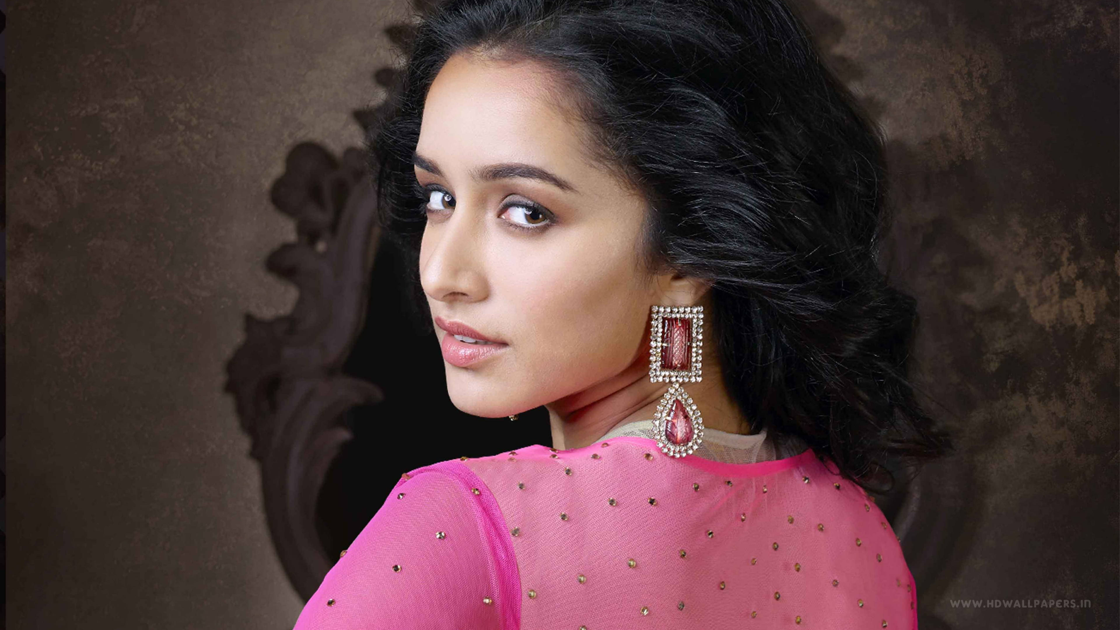 Shraddha Kapoor 桌布 壁紙 — 一位身穿鮮豔粉紅色裝飾上衣和華麗耳環的女性，在紋理棕色背景上越過肩膀凝視。