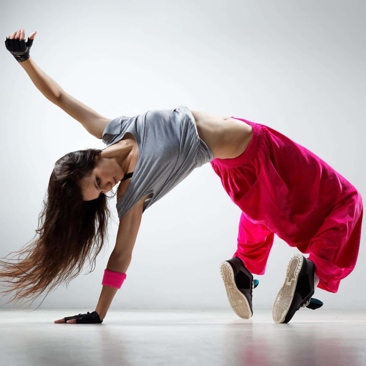 Hip Hop Dance 桌布 壁紙 — 一位身穿鮮豔粉紅色褲子和灰色上衣的女舞者，在純白色工作室背景前表演動態的霹靂舞動作。