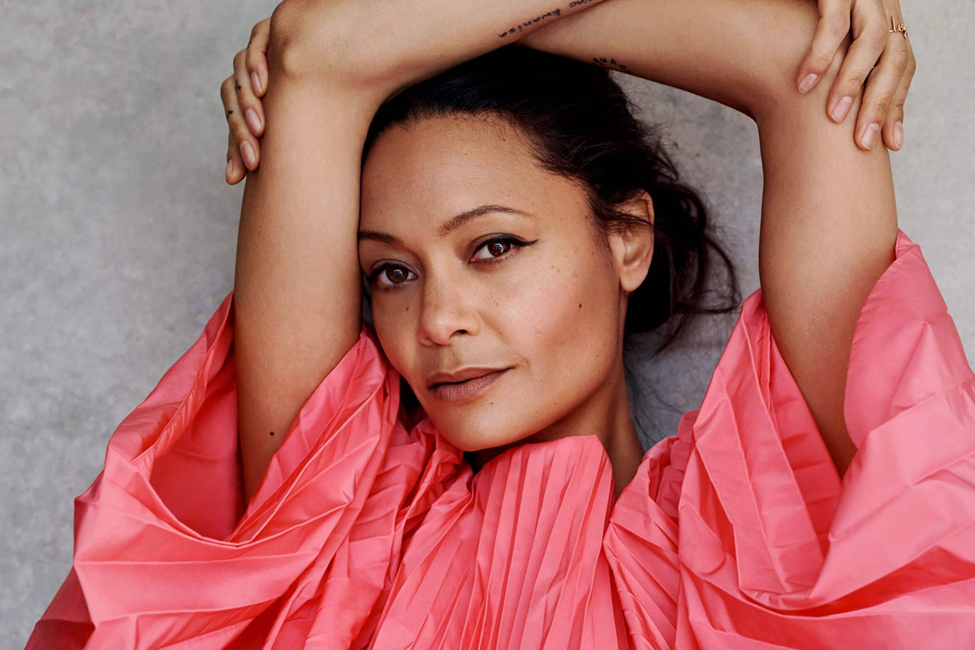 Actress HD 桌布 壁紙 — 一位身穿鮮豔粉紅色褶皺上衣的女性，在灰色中性背景前，雙臂彎曲舉過頭頂擺姿勢。