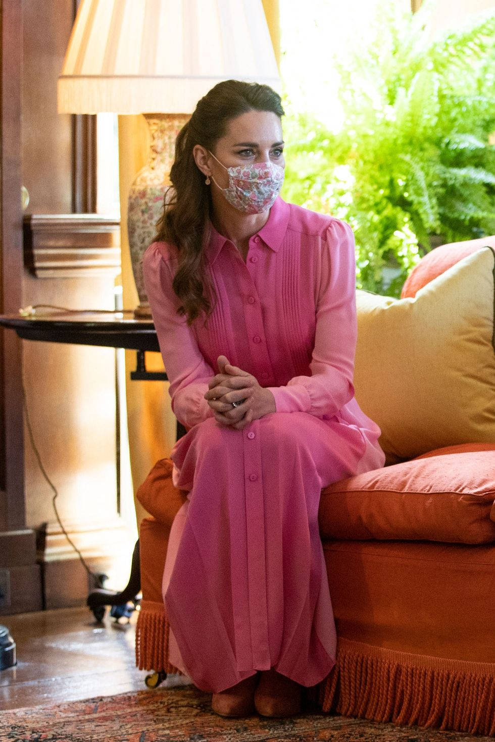 Kate Middleton 桌布 壁紙 — 一位身穿鮮豔粉紅色連衣裙的女性優雅地坐在橙色沙發上，室內光線溫暖，窗外可見茂密的綠色植物。