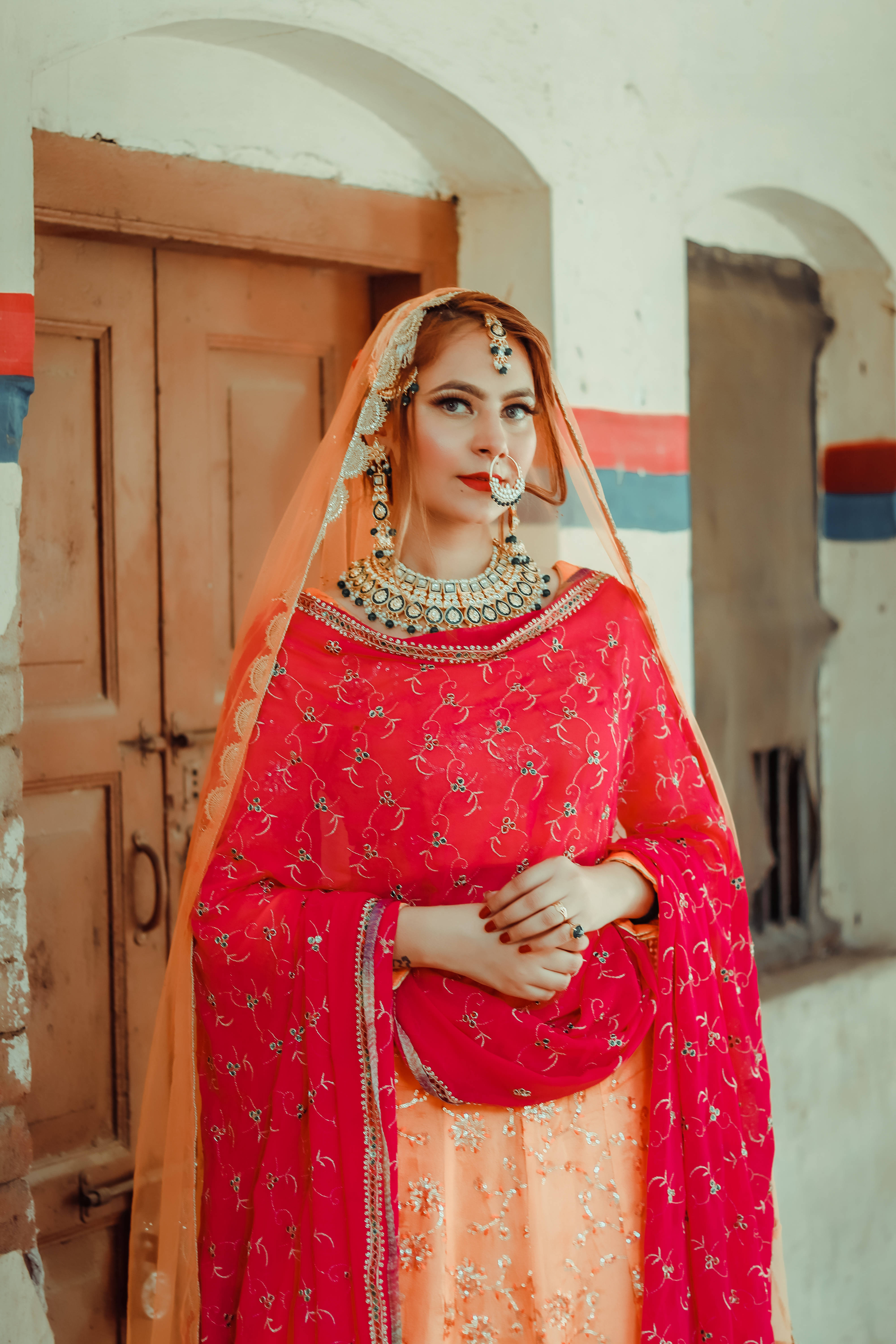 Indian 桌布 壁紙 — 一位身穿鮮豔紅桃色傳統服飾、帶有精緻刺繡和厚重珠寶的女性，優雅地站在質樸的門口。